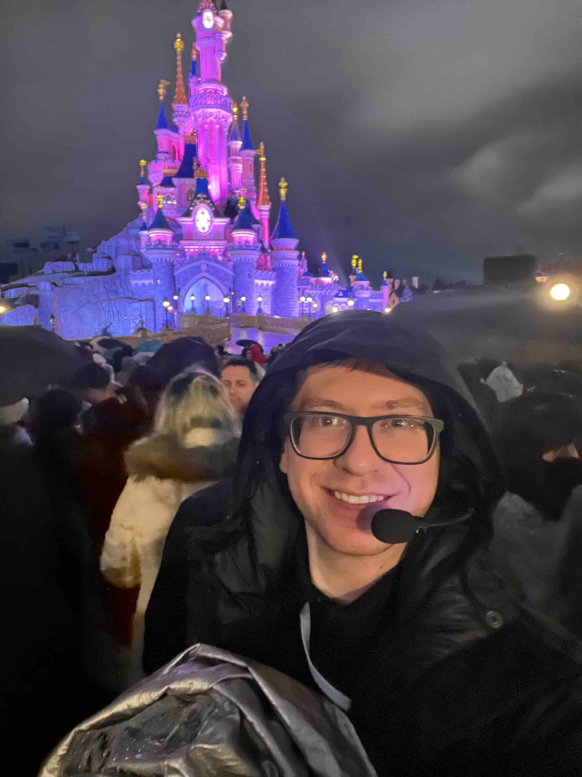 Man smiling in front of Sleeping Beauty Castle at Disneyland Paris, wearing glasses and a hooded jacket. Nighttime.