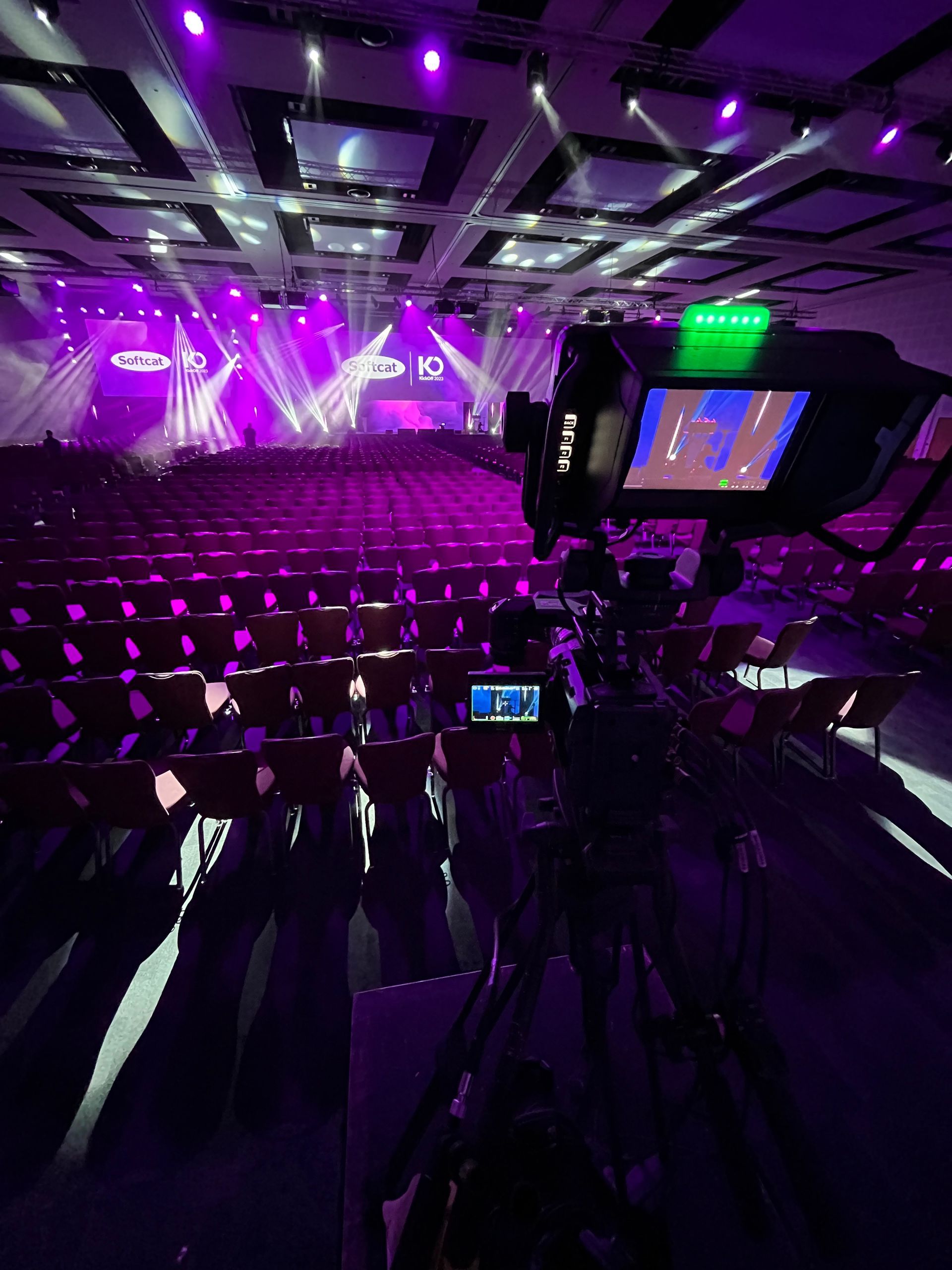 A professional video camera on a tripod in a large event hall, stage lit with purple, rows of seats.