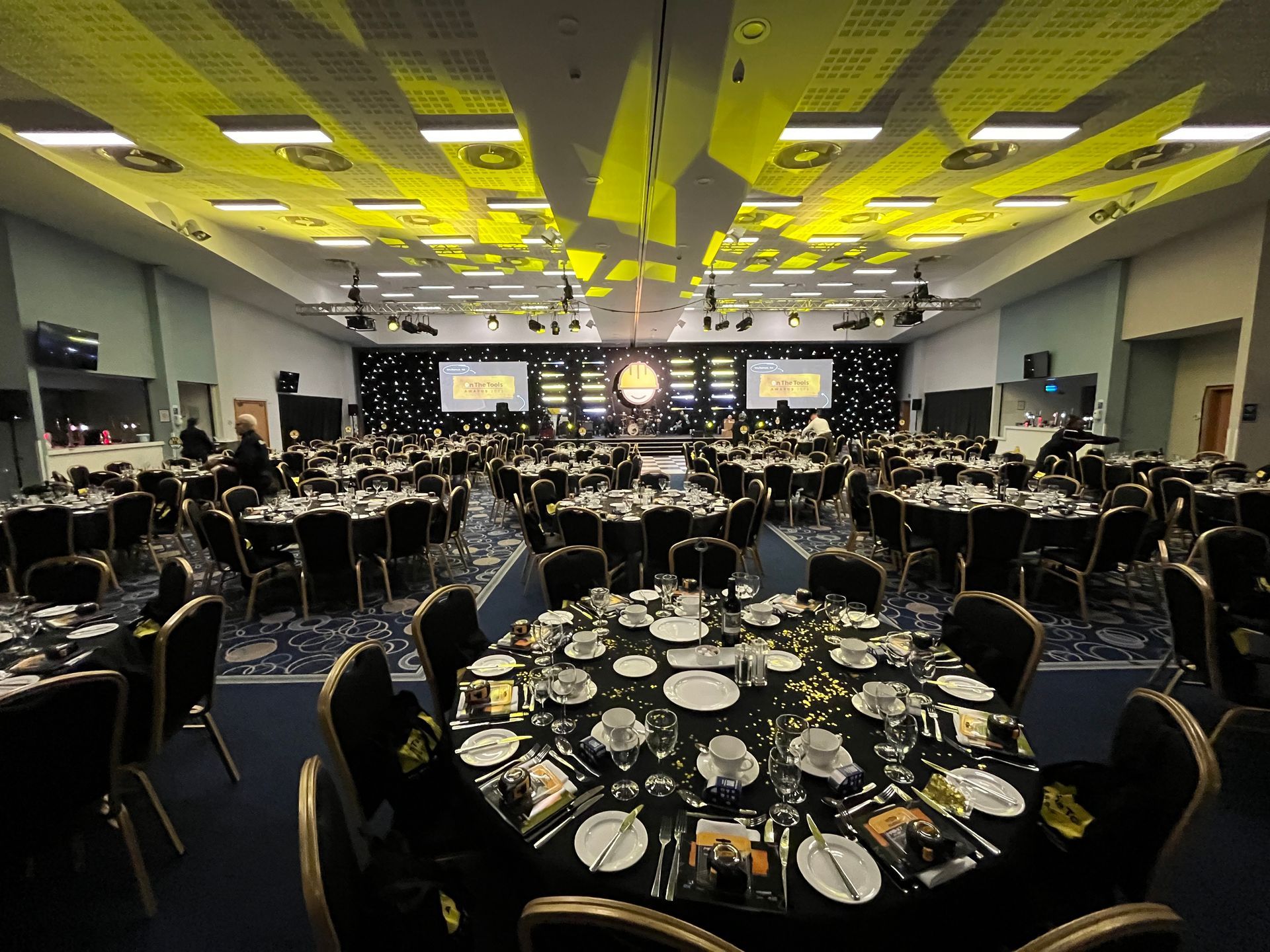 Formal event hall with round tables, black tablecloths, and stage in the distance. Yellow lighting and decor.
