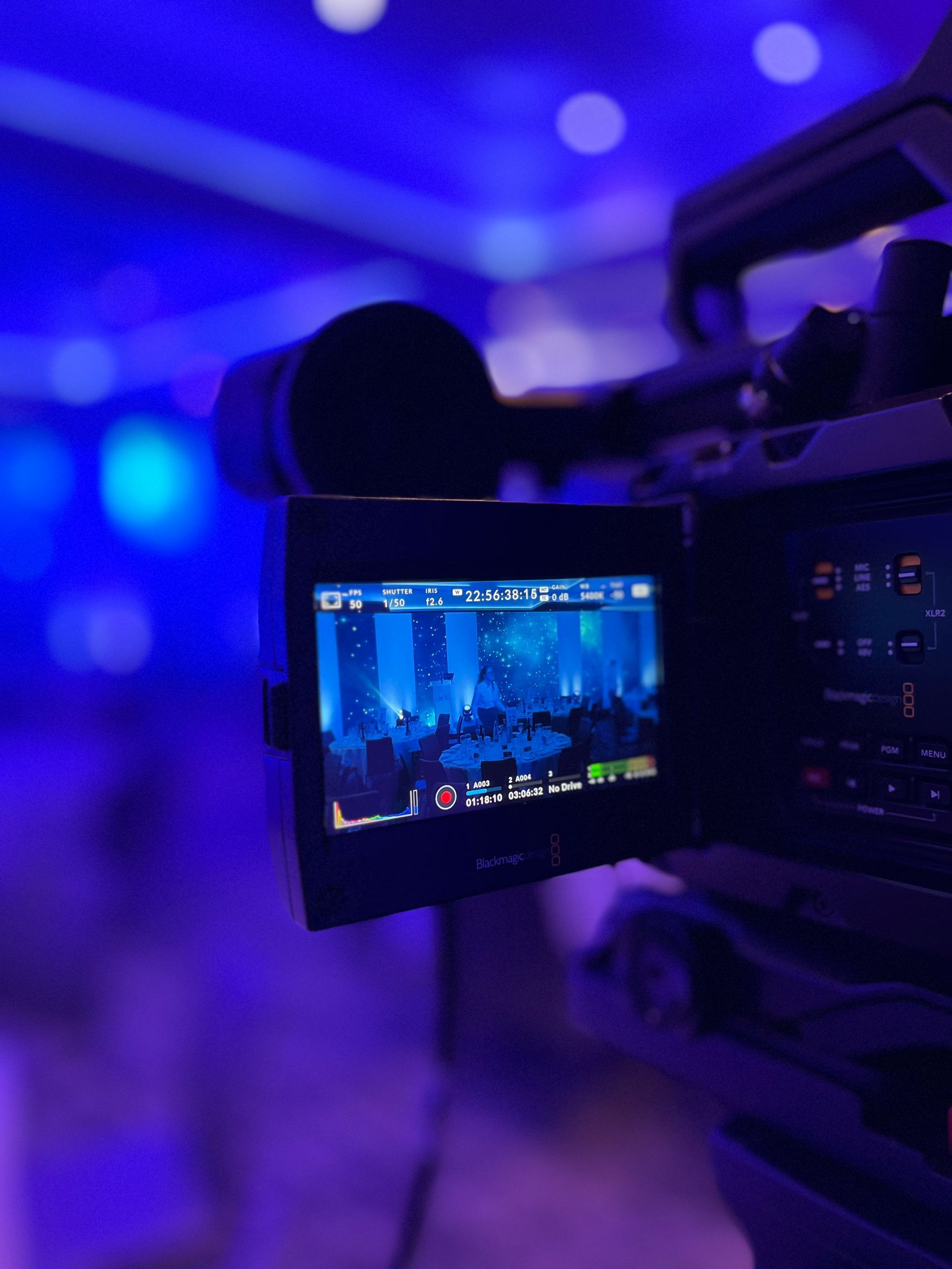 Video camera filming a scene with a blue-lit backdrop, screen showing the image being recorded.