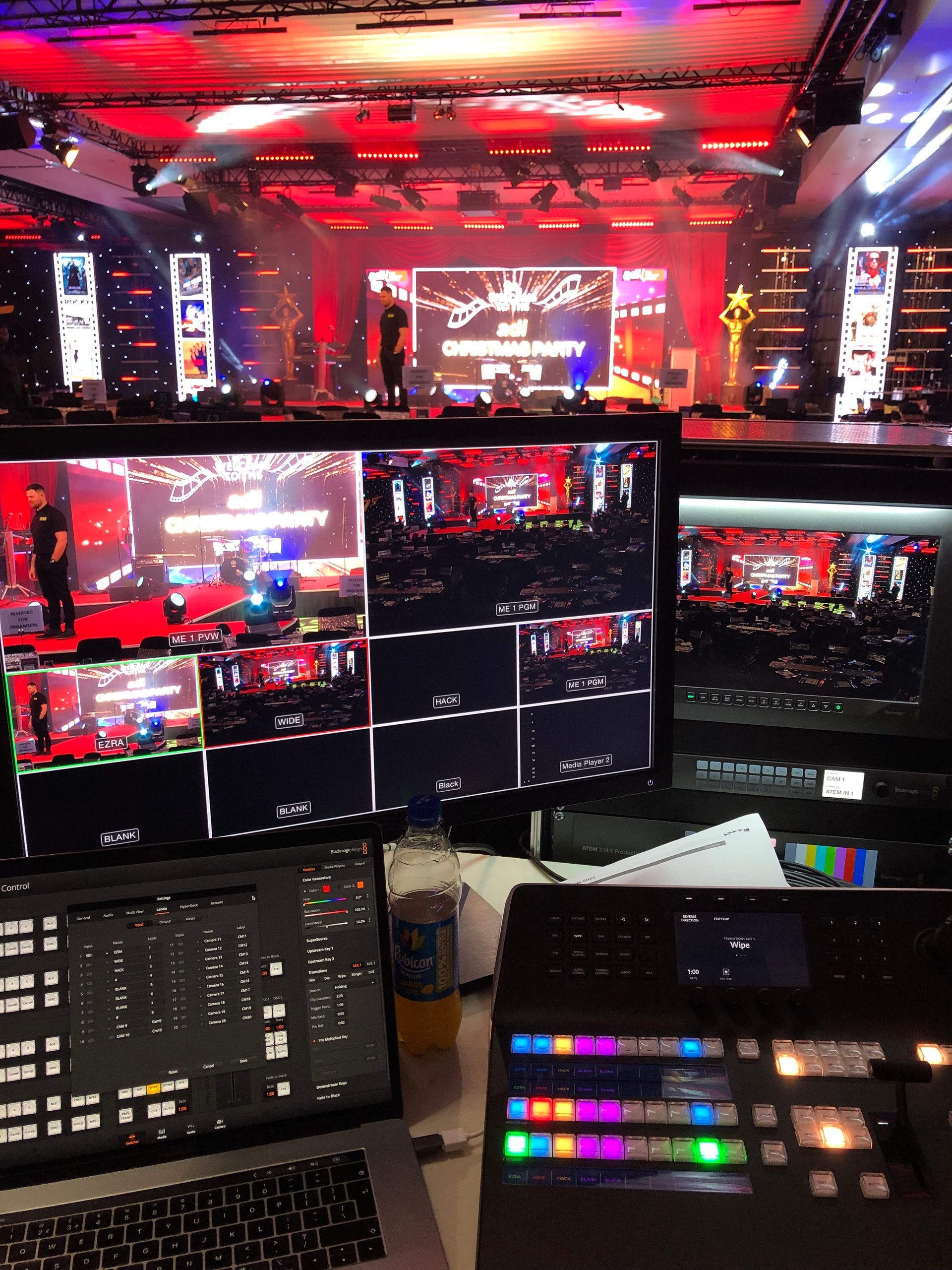 Production control room with monitors displaying stage view and event feed. Red lighting and stage with people present.