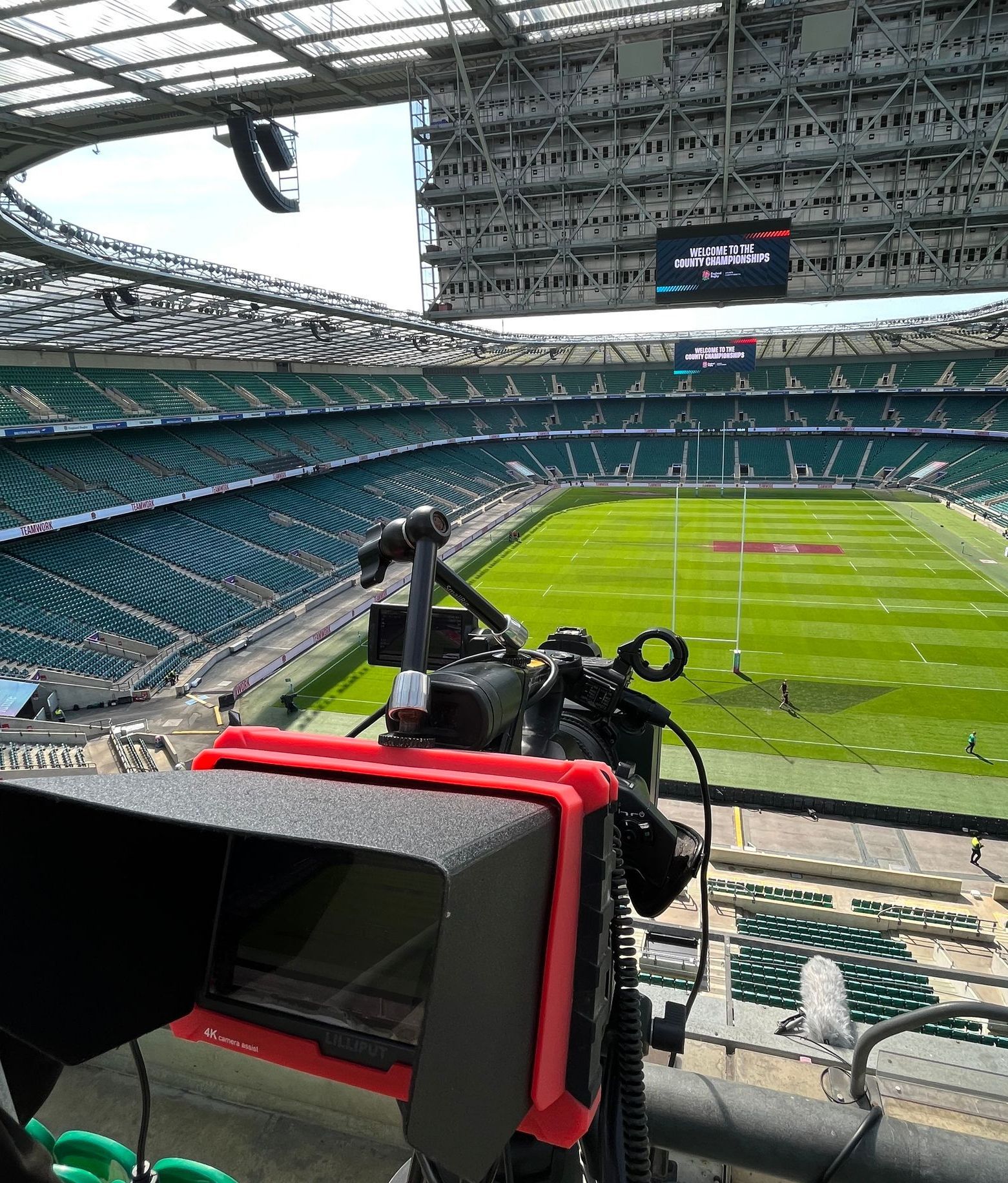 A camera on a tripod overlooking a rugby field in a stadium with green seating and a large scoreboard.