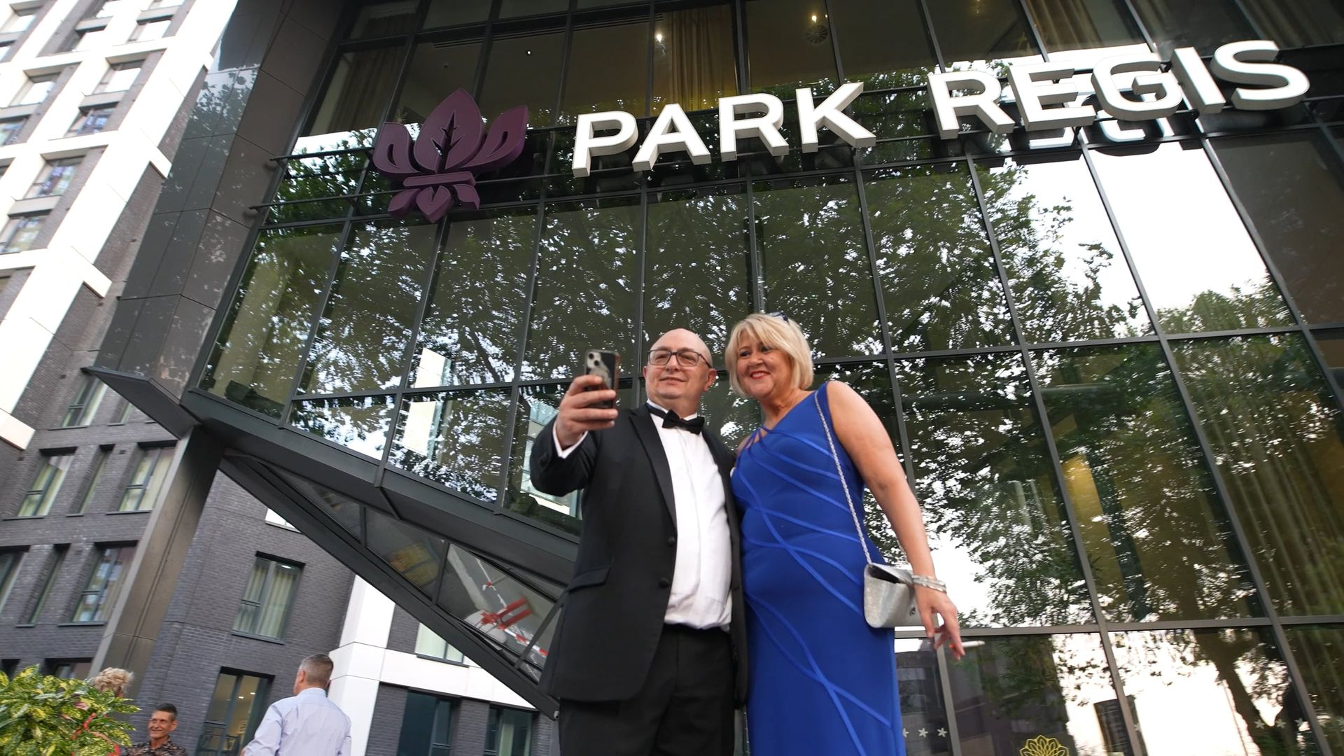 Man and woman taking selfie in front of the Park Regis hotel. Both are dressed formally.