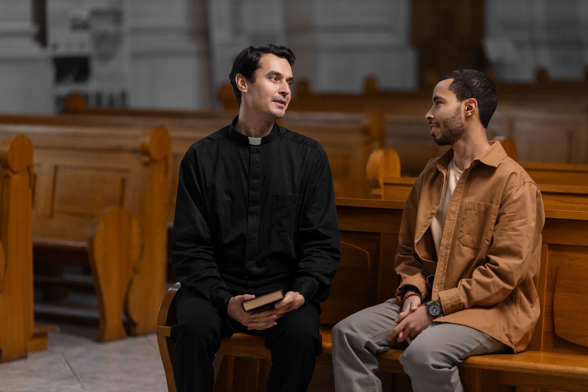 Two men sit talking on church pews in a wooden chapel.