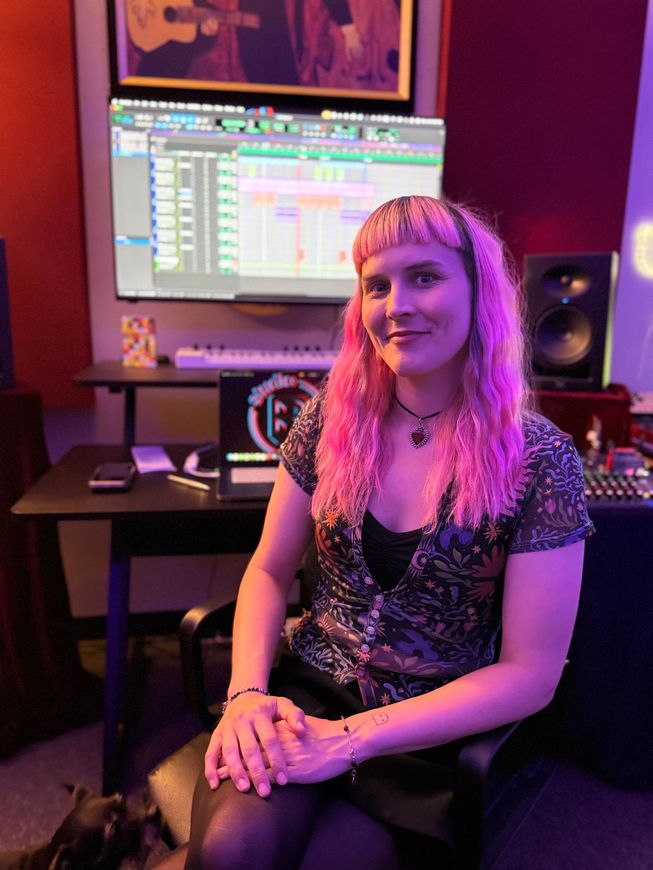 Person seated in a neon-lit studio, smiling in front of a computer monitor and audio speaker.