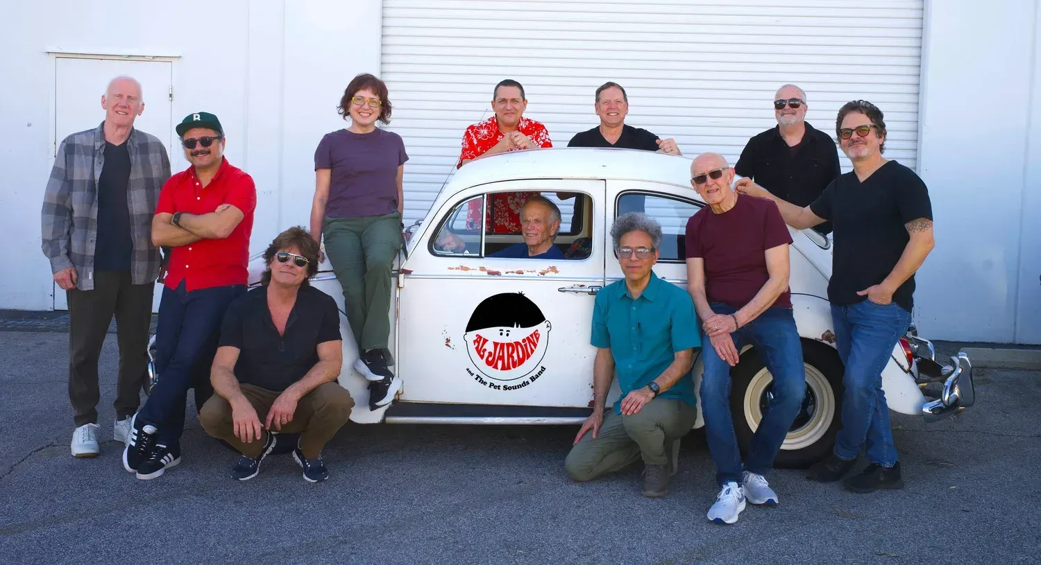 Group posing around a small white car in front of a garage door, smiling and kneeling together.