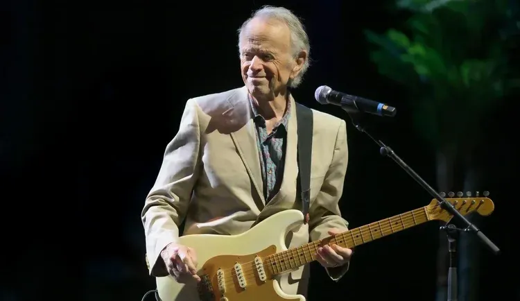 Musician playing a cream electric guitar onstage beside a microphone, wearing a light suit under stage lights
