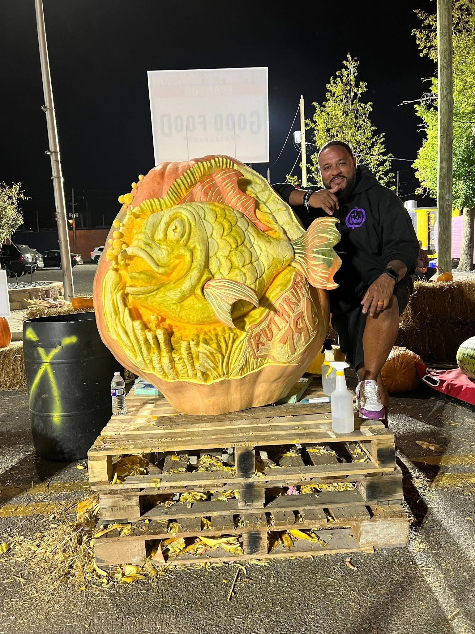 Man holding a large carved pumpkin with a face, lit by lanterns on a nighttime outdoor stage