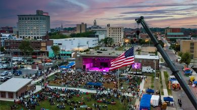 Crowd at an outdoor concert at dusk with a large American flag and a bright magenta stage in the city center