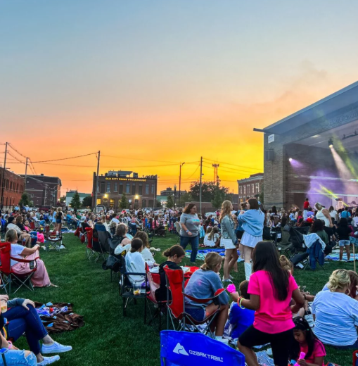 Crowd seated on a grassy lawn at an outdoor concert at sunset with stage lights glowing in the distance