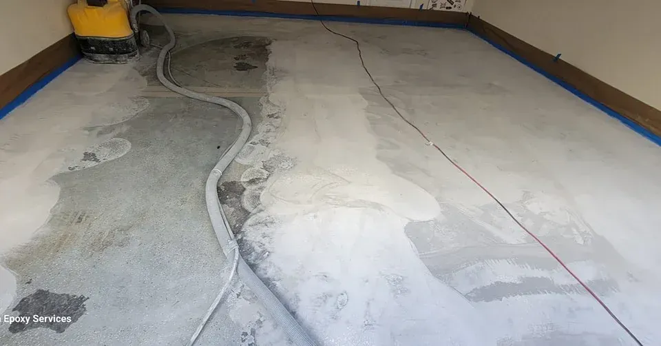 Garage floor being cleaned by a machine. Blue tape lines edges. Light gray concrete floor with wet and dry patches.