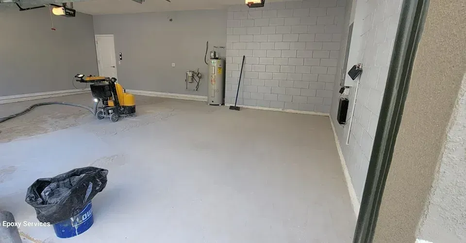 Garage interior: concrete floor being resurfaced with a grinding machine; gray walls, a water heater, a trash bag.