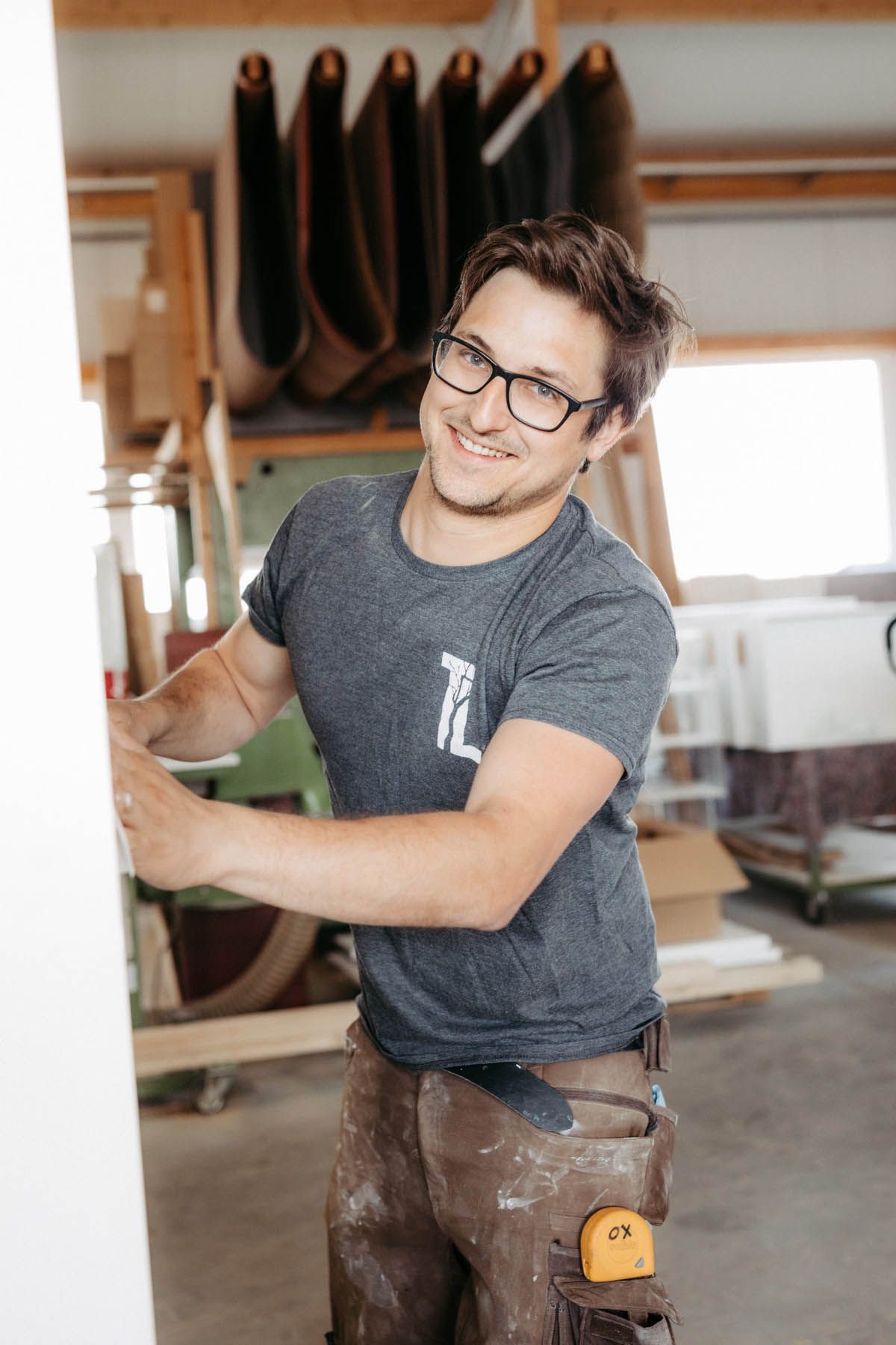Lächelnder Zimmermann in einer Werkstatt, der Holz hält; trägt Brille, graues T-Shirt und Arbeitshose.