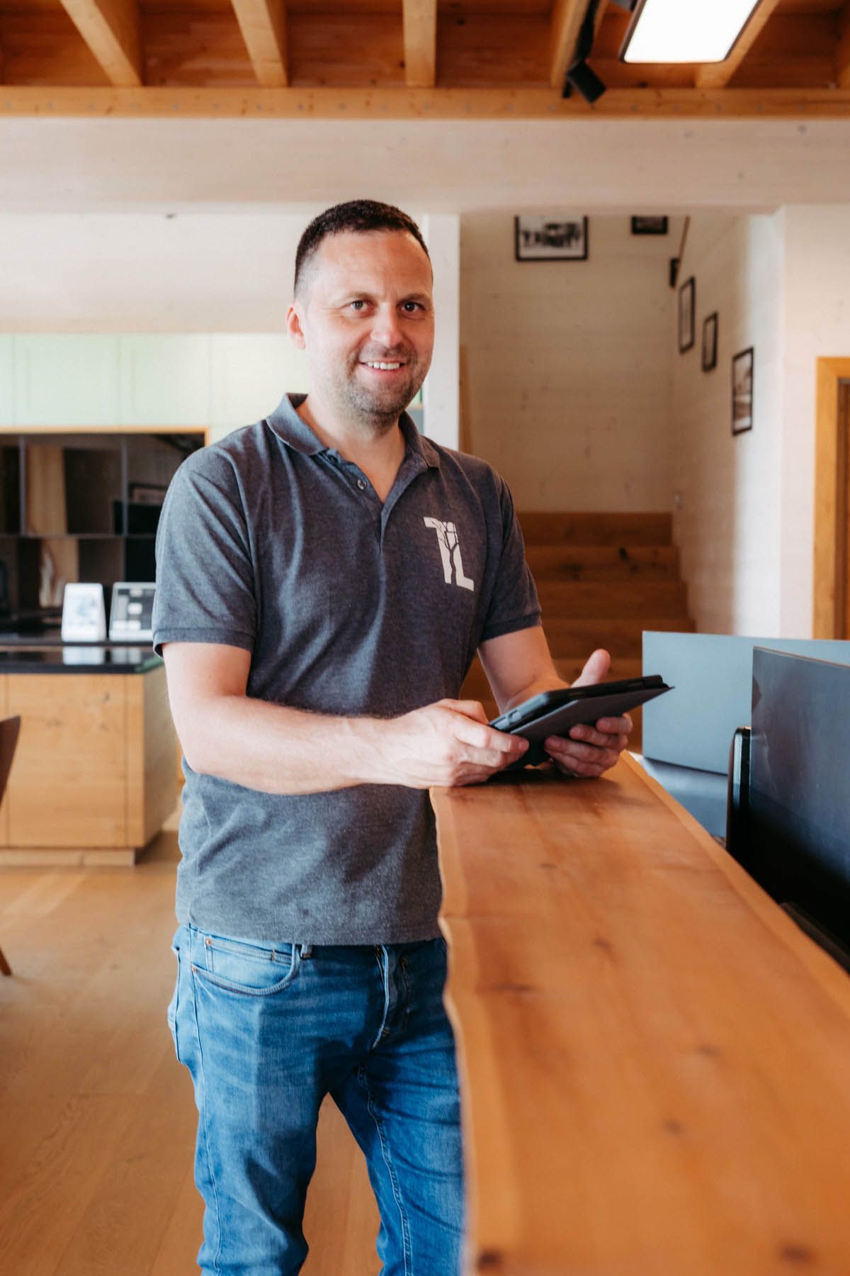 Mann hält Tablet, lächelt, lehnt an einer Holztheke, trägt ein graues Poloshirt und Jeans.