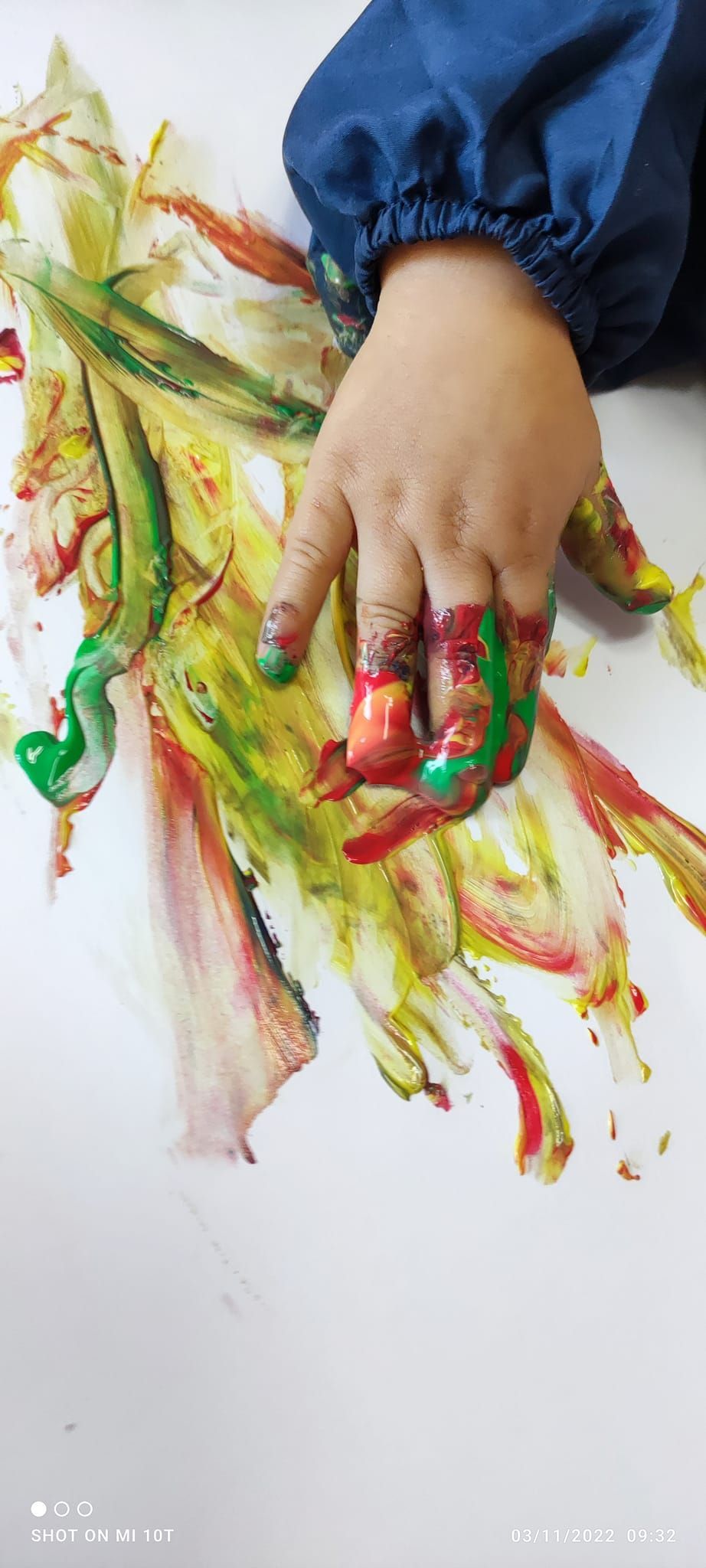Mano con le dita macchiate di pittura che traccia tratti colorati su carta bianca, manica di una maglietta blu visibile