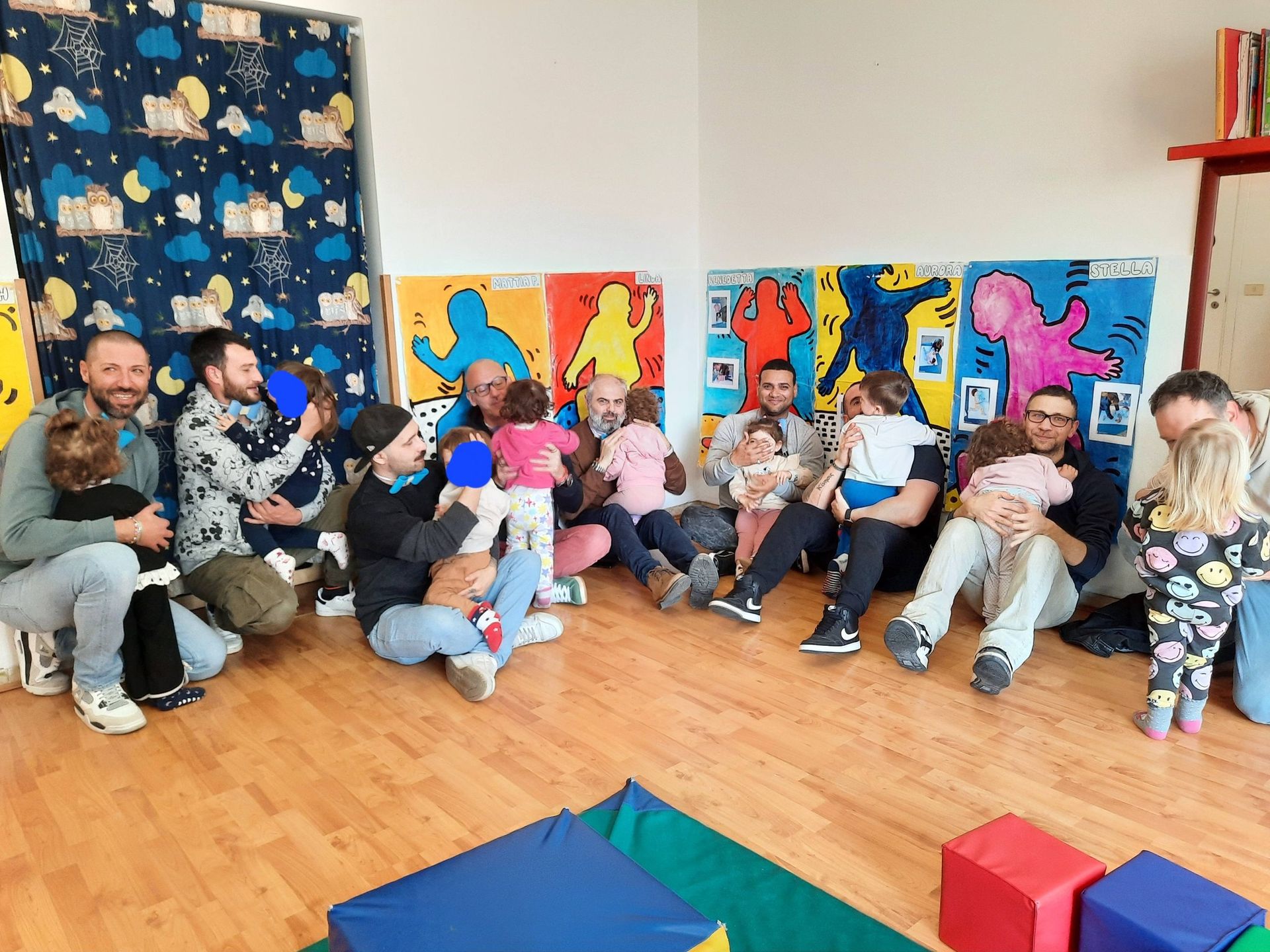 Gruppo di bambini seduti sul pavimento in un'aula colorata, con pannelli artistici e giocattoli nelle vicinanze.