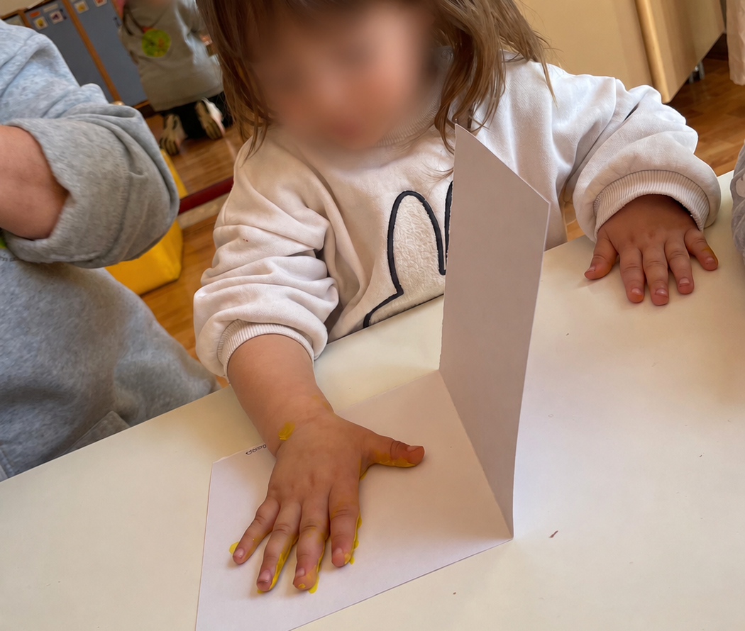 Un bambino piega la carta a un tavolo con un cartoncino bianco e una ciotola verde in un'aula scolastica.