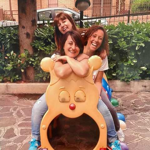 Tre donne posano su una colorata giostrina a forma di cane in un parco giochi allestito in un giardino.