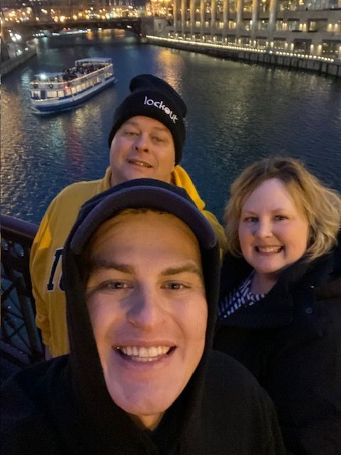 Three people smiling on a nighttime boat ride by city lights and water
