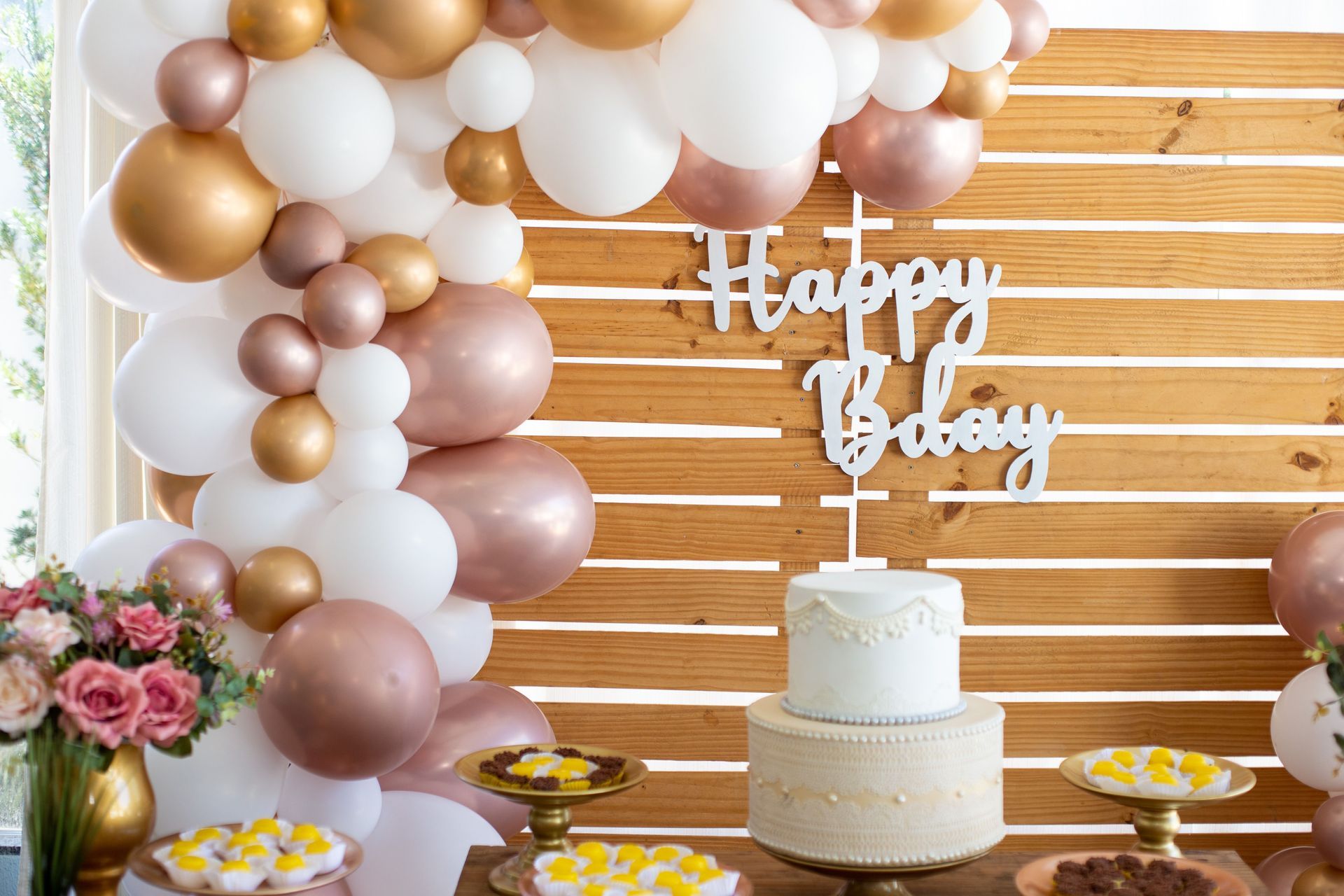 Birthday dessert table with white, gold, and blush balloons, cake, flowers, and a “Happy Birthday” sign