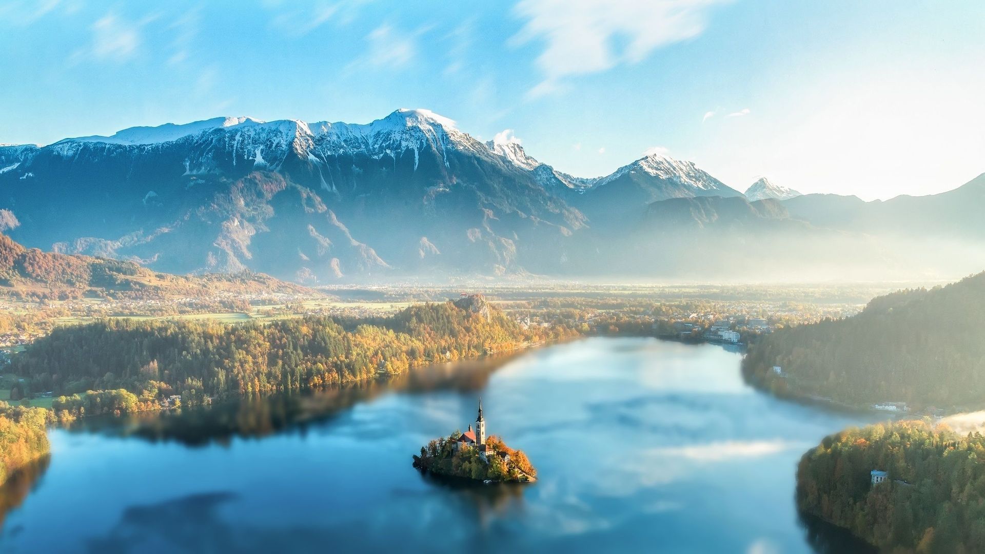 Snowy mountains rise above a misty blue lake with a small tree-covered island in the foreground.