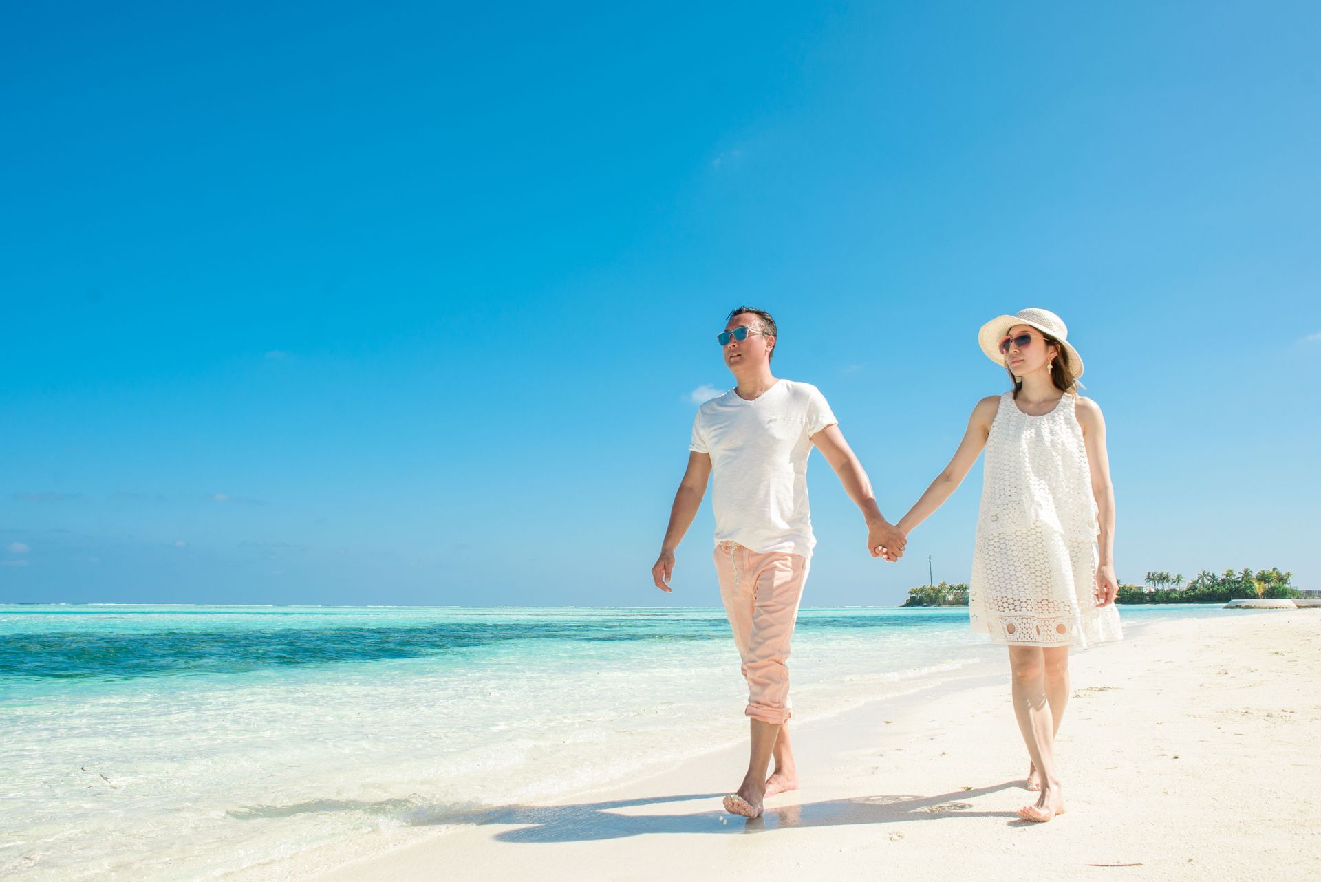 Two people in white walking hand in hand on a sunny beach with turquoise water and a blue sky