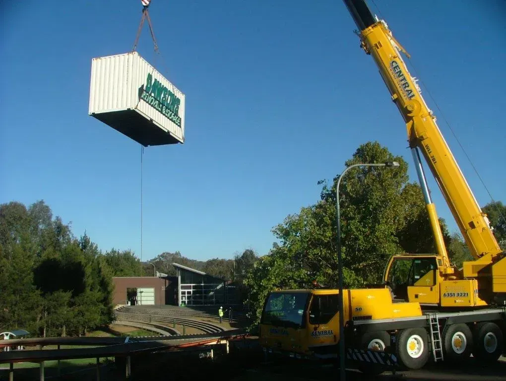 A Yellow Crane Lifting a White Shipping Container With Green Logo — Dawson's Removals In Kelso, NSW