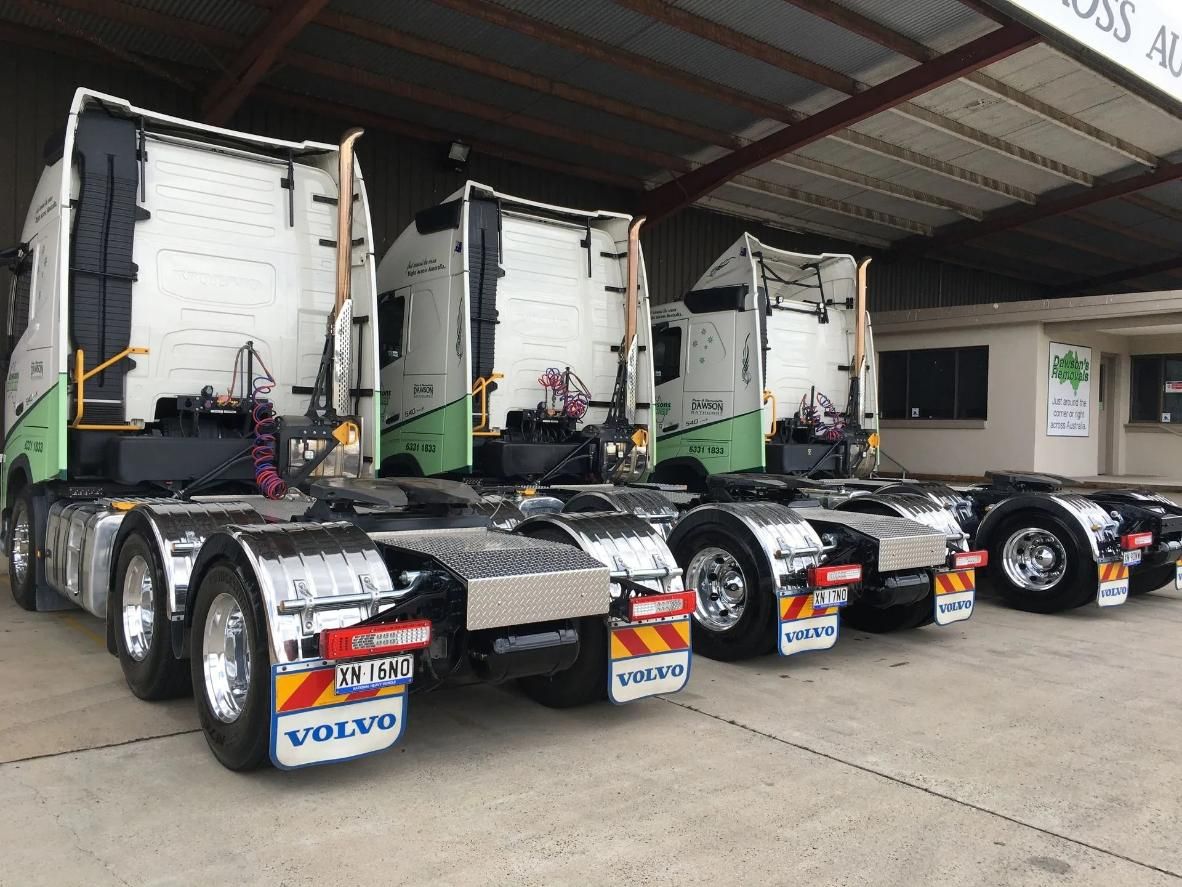 Three parked Volvo semi-trucks, seen from the rear, under a shed roof. Silver, white, and green color scheme.