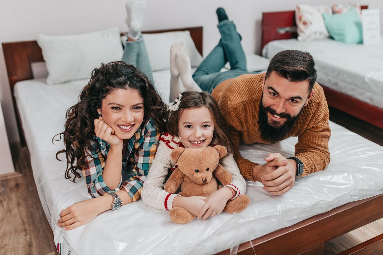 Three people lying on a bed, smiling and holding a teddy bear, with suitcases in the background.