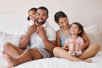 Family sitting together on a bed, smiling and hugging in a bright bedroom.