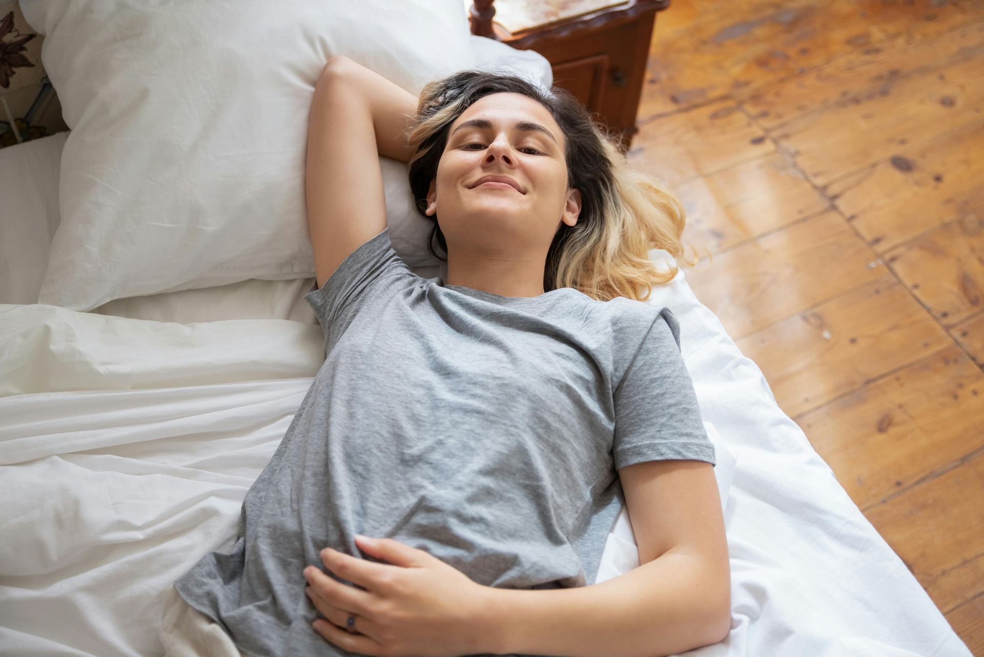 Person lying on a bed with eyes closed and hand on stomach, wearing a gray shirt in a bright room.