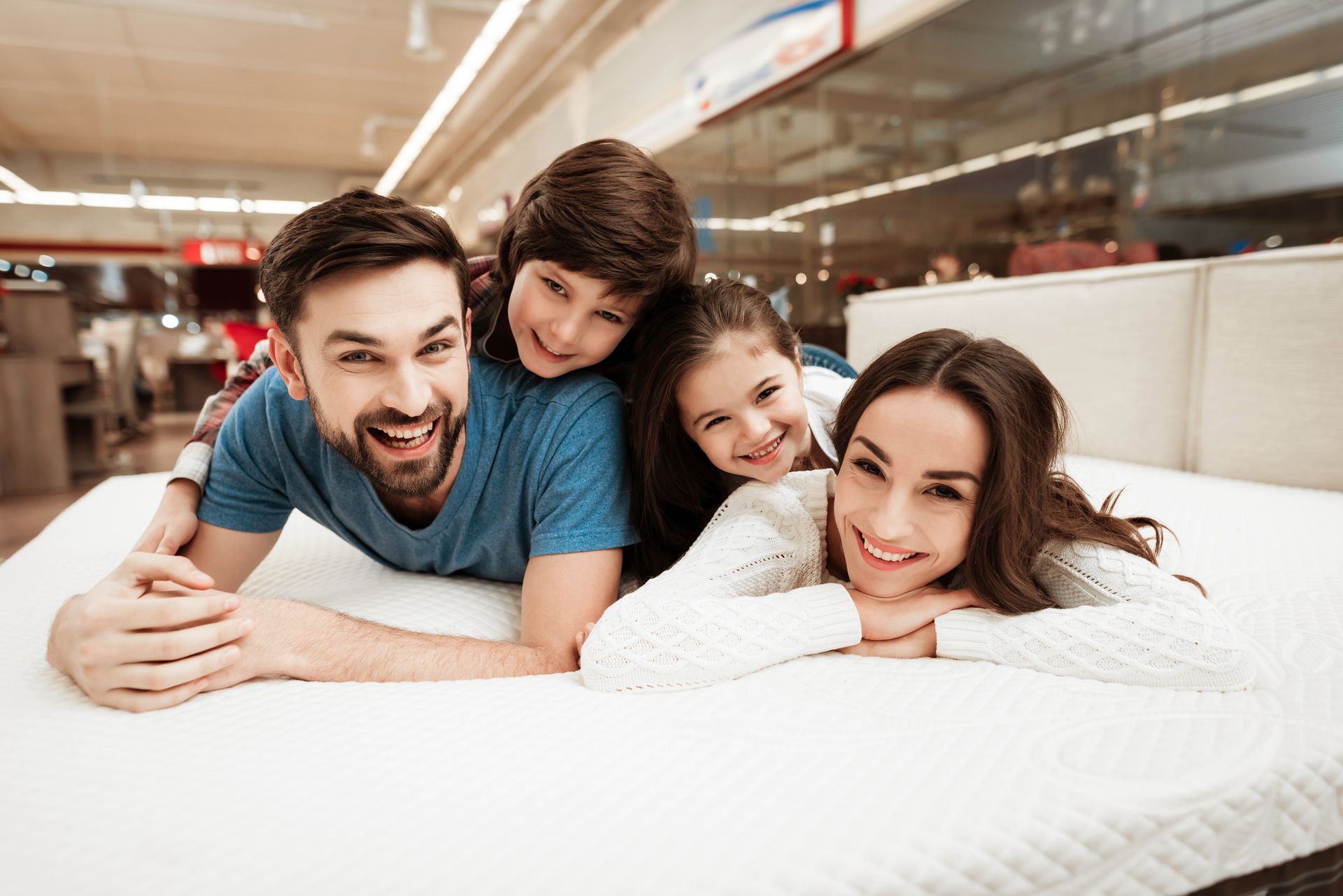Family smiling on a bed in a bright bedroom, with two adults and two children leaning close together.
