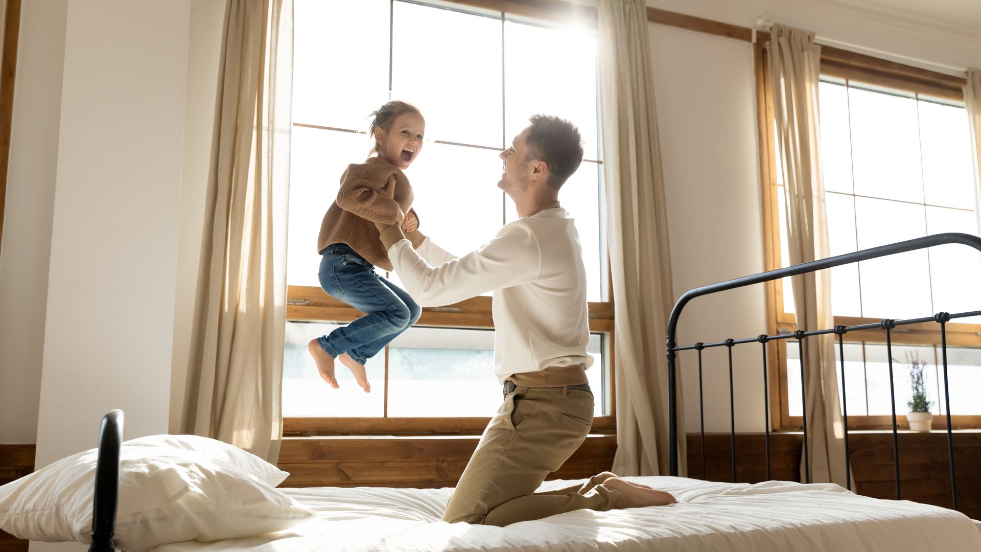 Parent lifting smiling child on a bed in a bright bedroom near large windows.