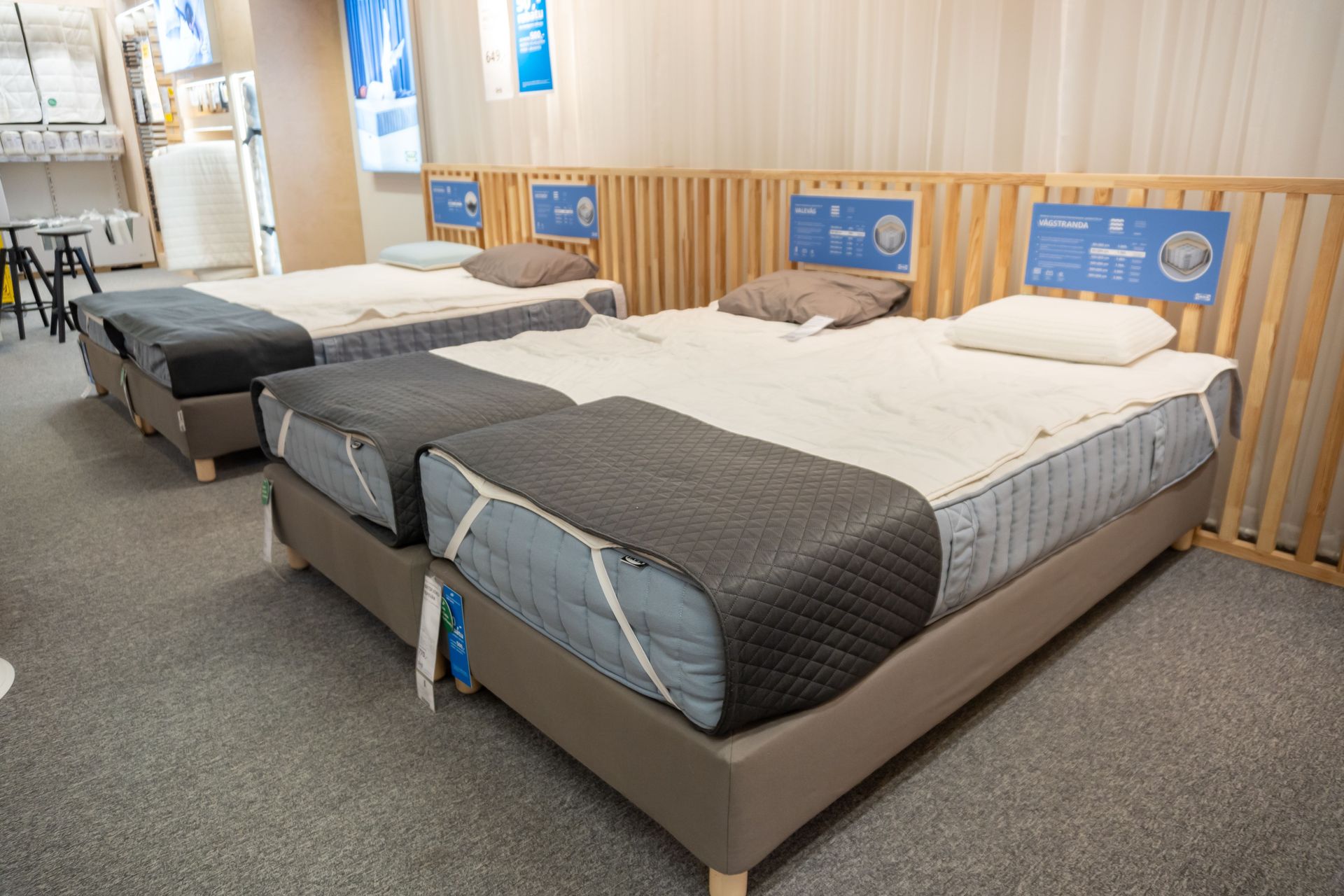 Three neatly made beds in a showroom with blue headboard signs and gray carpet.