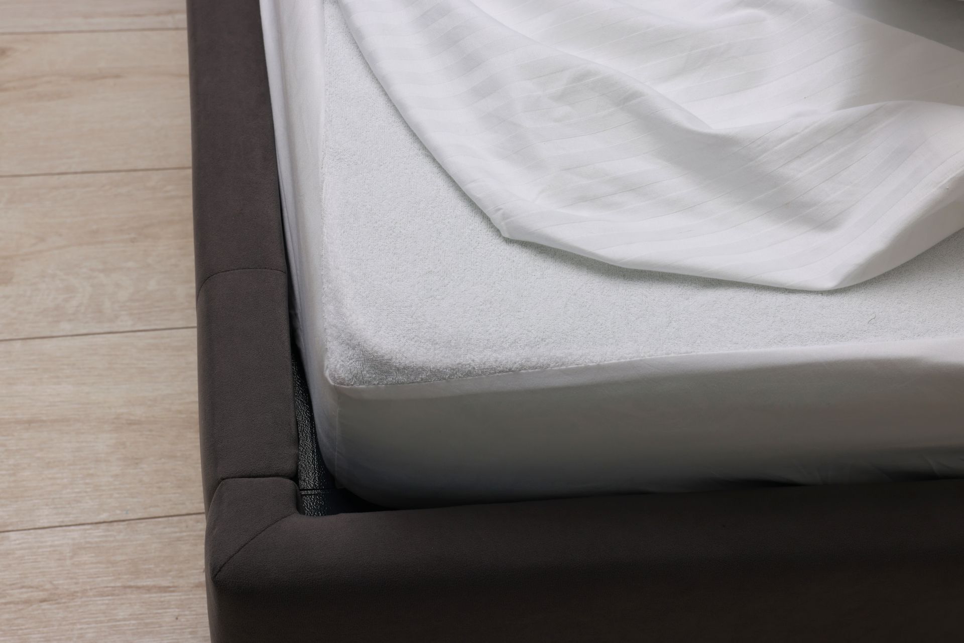 Close-up of a bed corner with a white sheet and dark upholstered frame on a wooden floor.