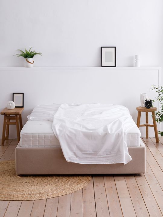 Minimal white bedroom with a bed, wood stools, wall shelf, and woven rug in a bright, airy space.