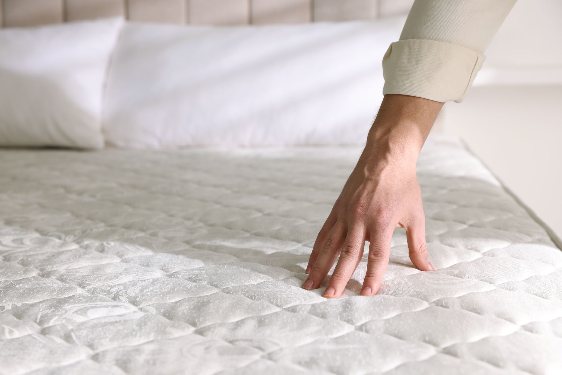 Hand checking a white mattress in a bright bedroom.