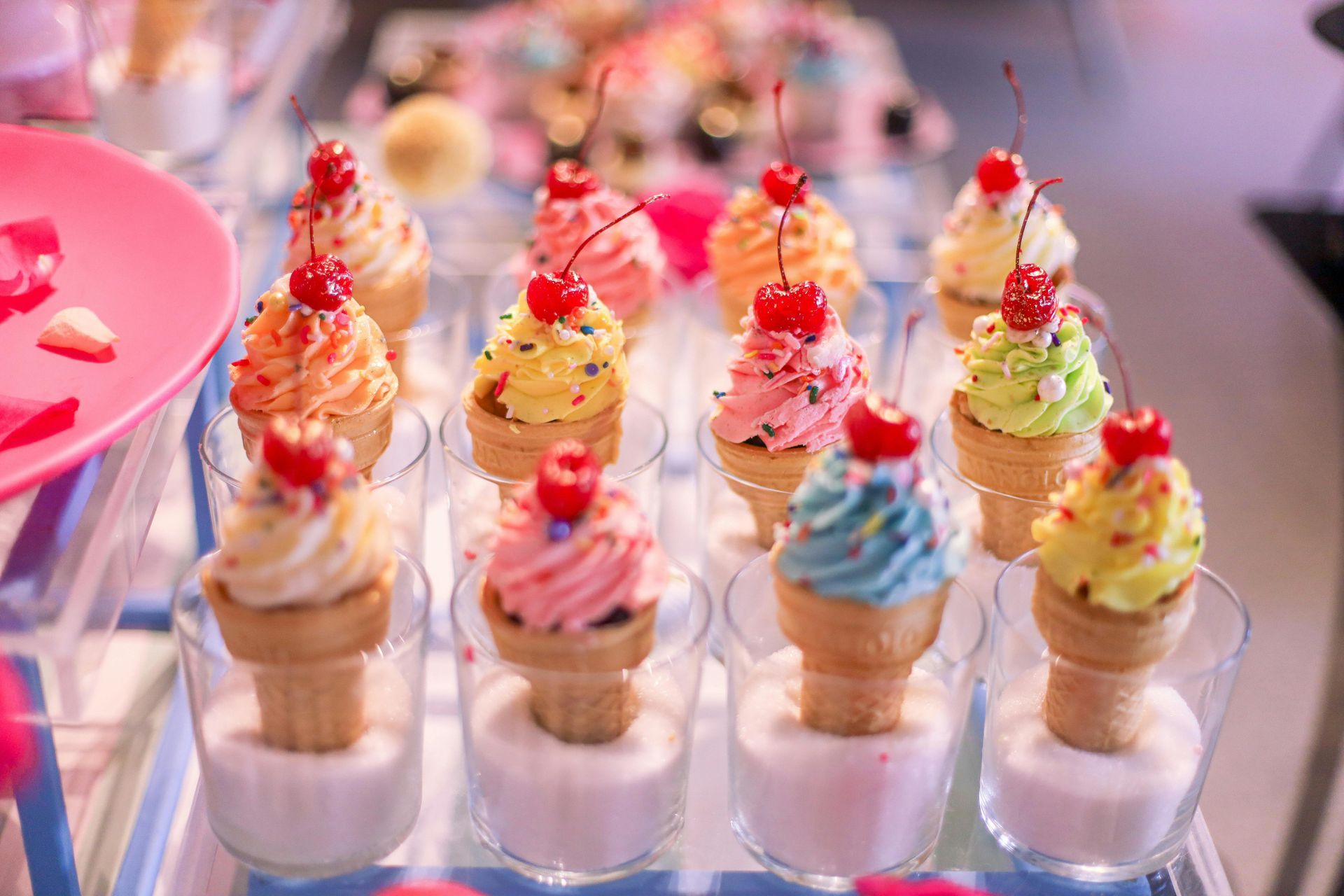 Small ice cream cone-shaped desserts with colorful frosting and a cherry on top, displayed in clear cups filled with sugar.