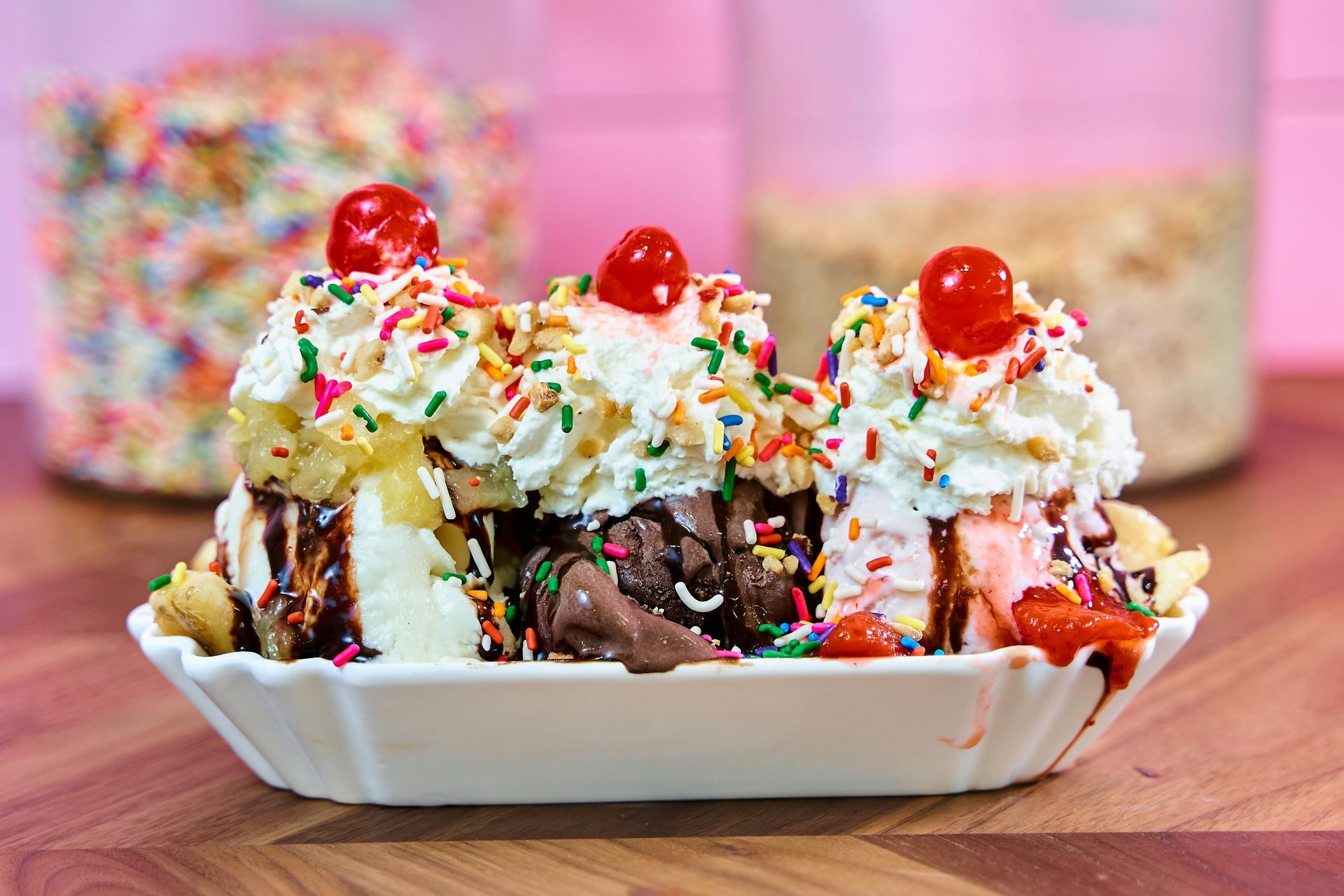 A banana split with three scoops of ice cream, whipped cream, chocolate syrup, rainbow sprinkles, and three red cherries.