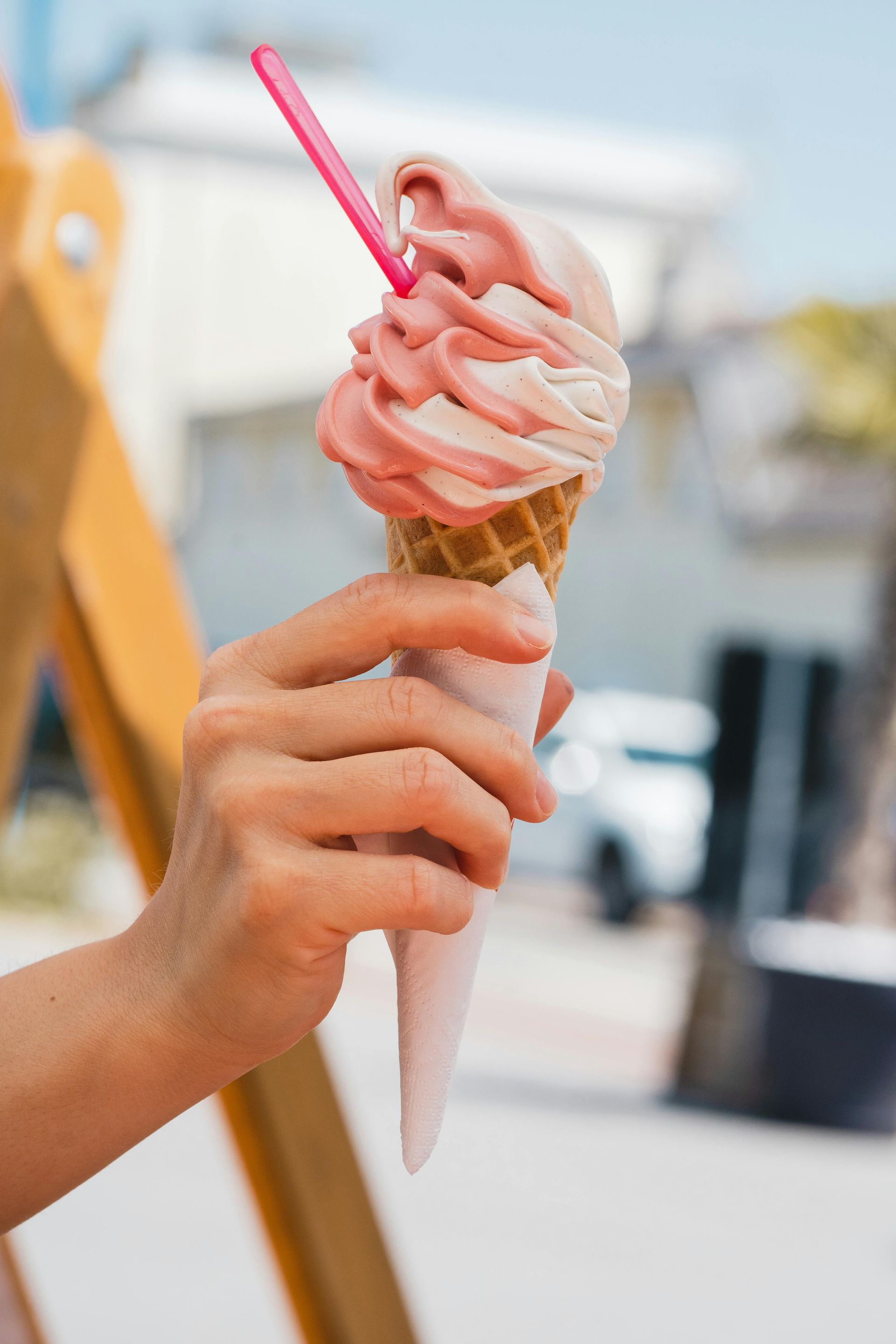 A hand holds a swirled pink and white soft-serve ice cream cone with a pink plastic spoon in a sunny outdoor setting.