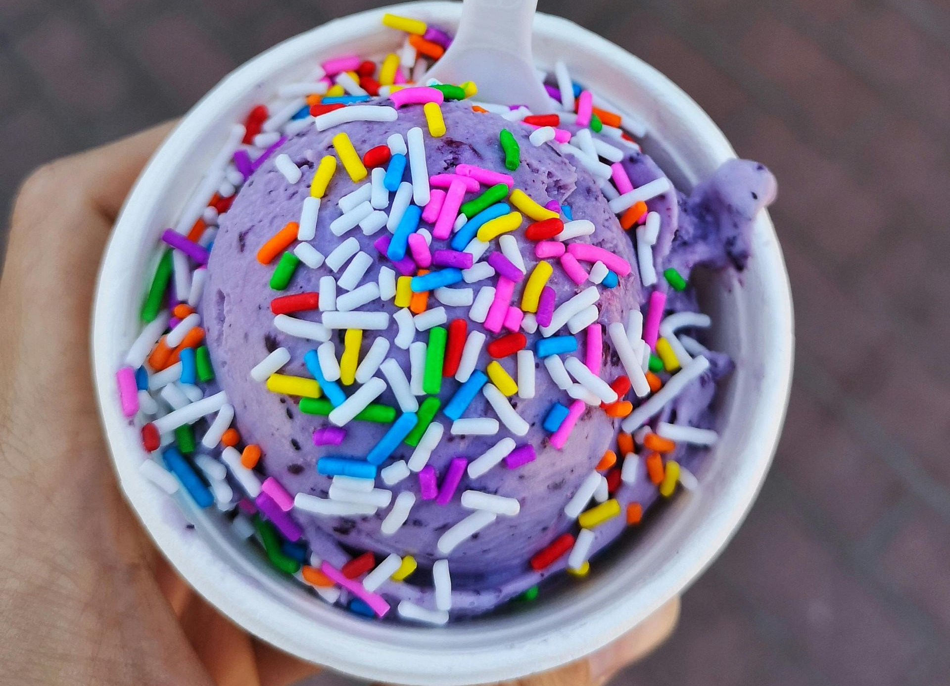 A person holds a white cup of purple ice cream topped with colorful sprinkles and a plastic spoon.