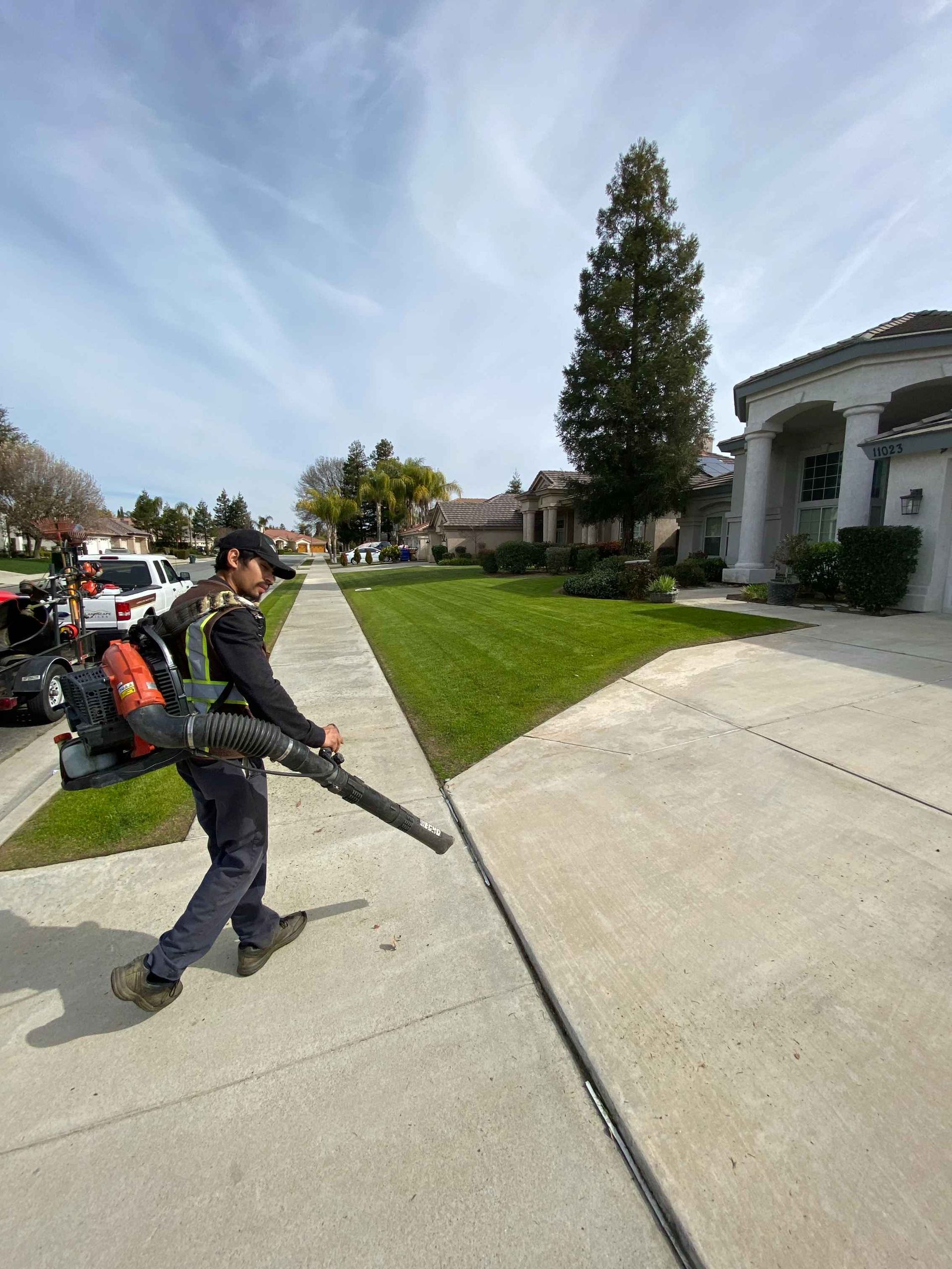 Man blowing debris from a sidewalk next to a green lawn. Residential neighborhood with cloudy sky.