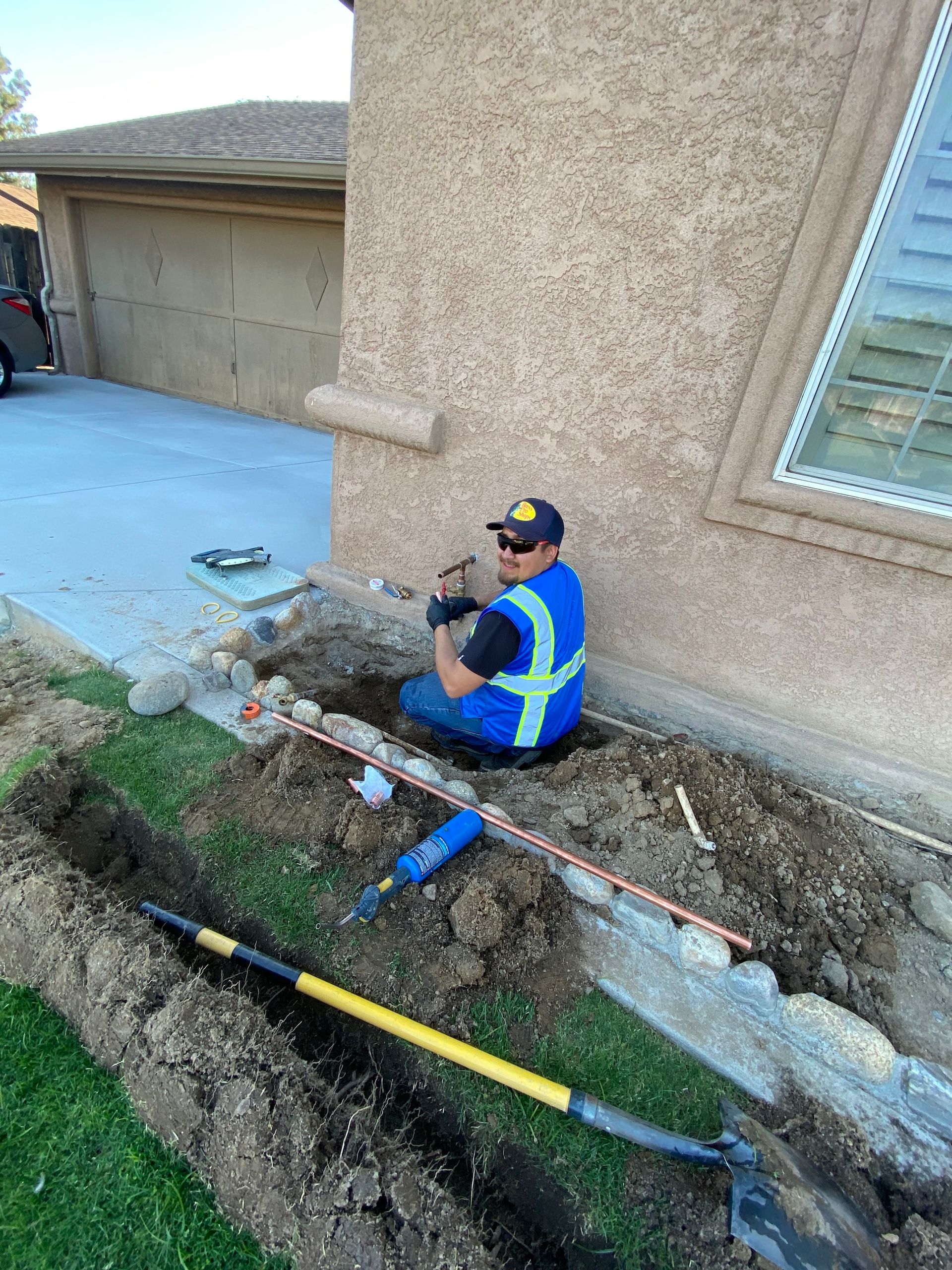 Man in vest working on pipes in trench near house.