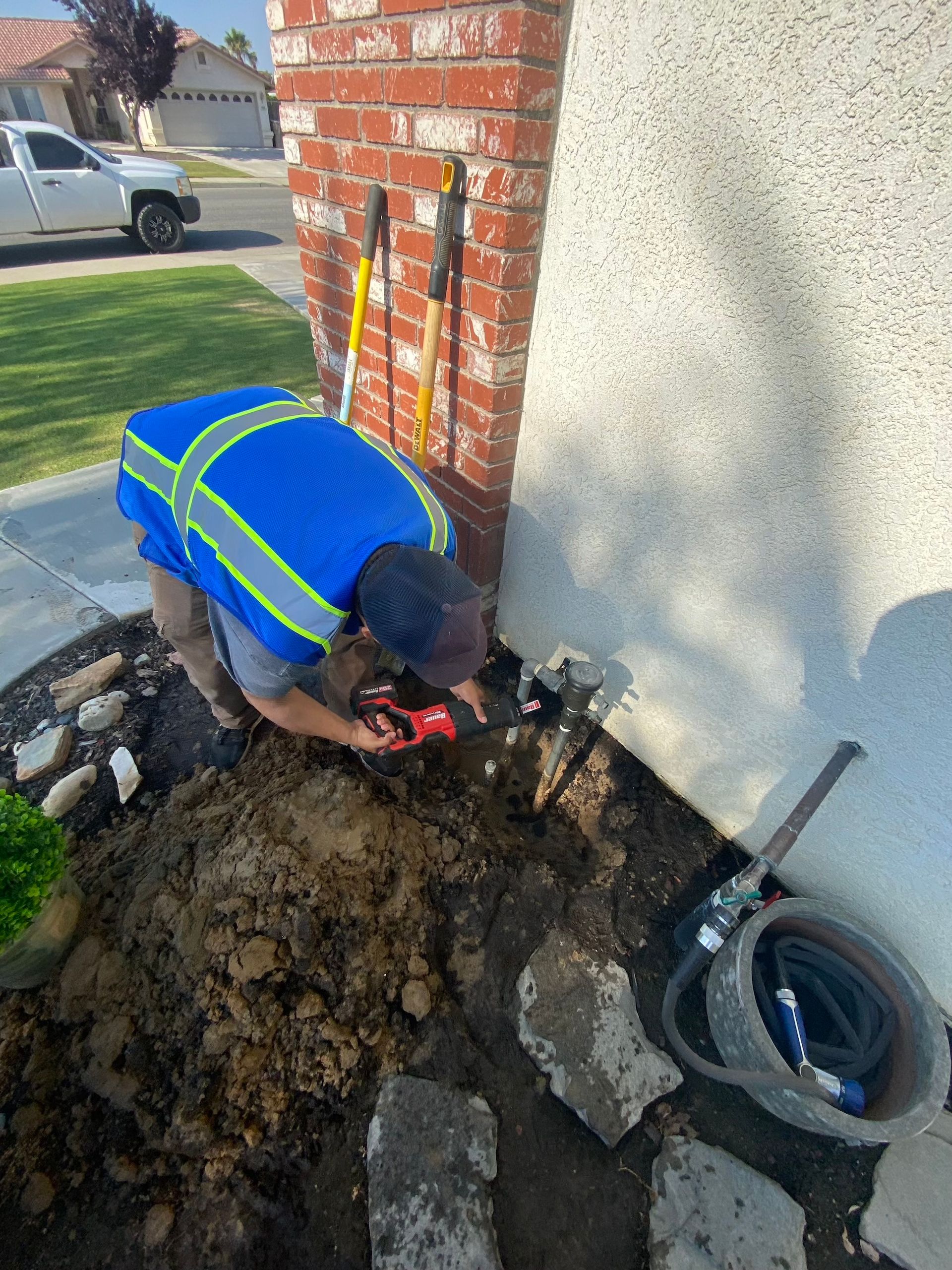 Person in blue vest working on ground near a brick pillar, tools nearby, hose and bucket visible.