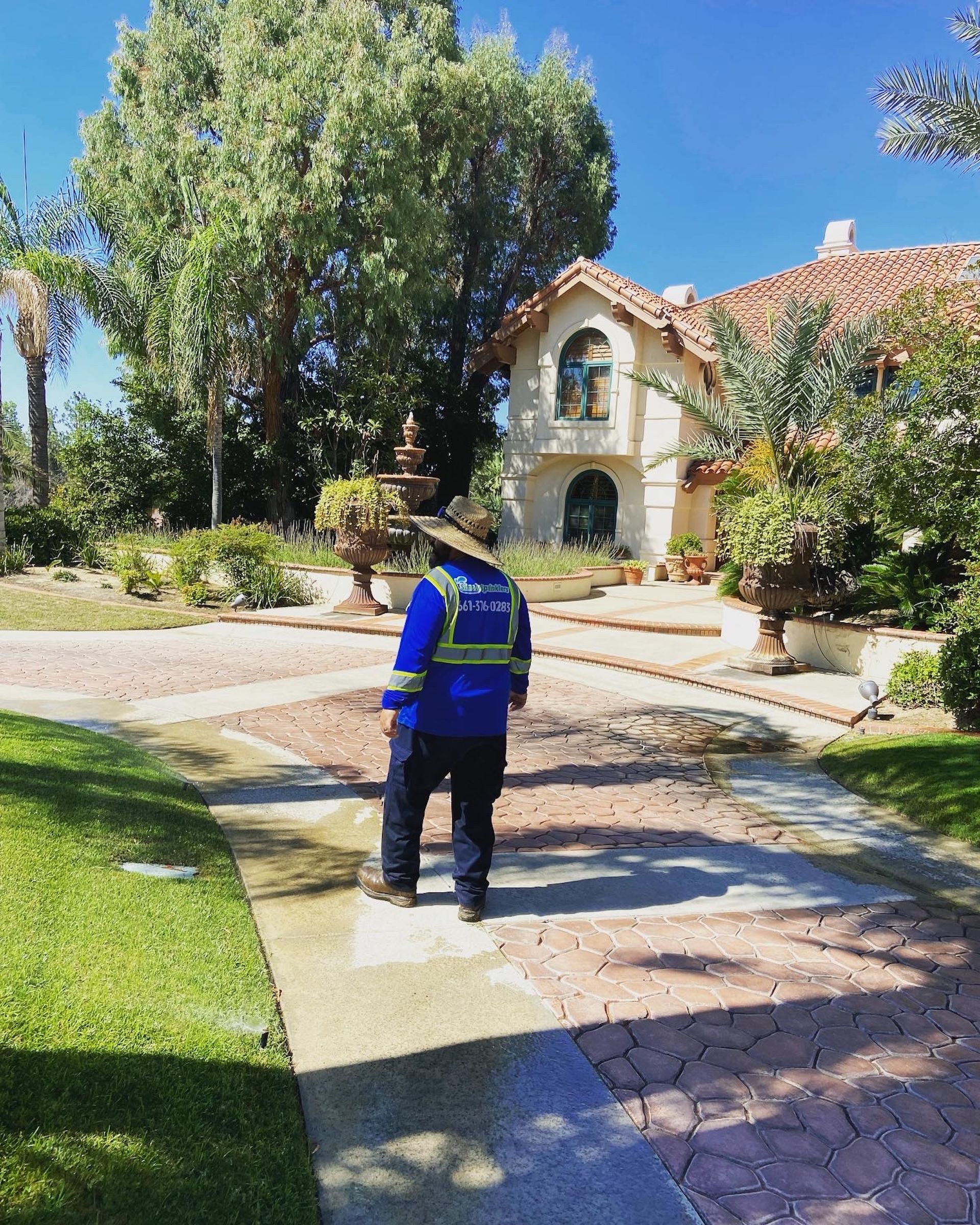 Person in blue uniform standing on brick pathway in front of a house. Sunny day, trees, lawn.