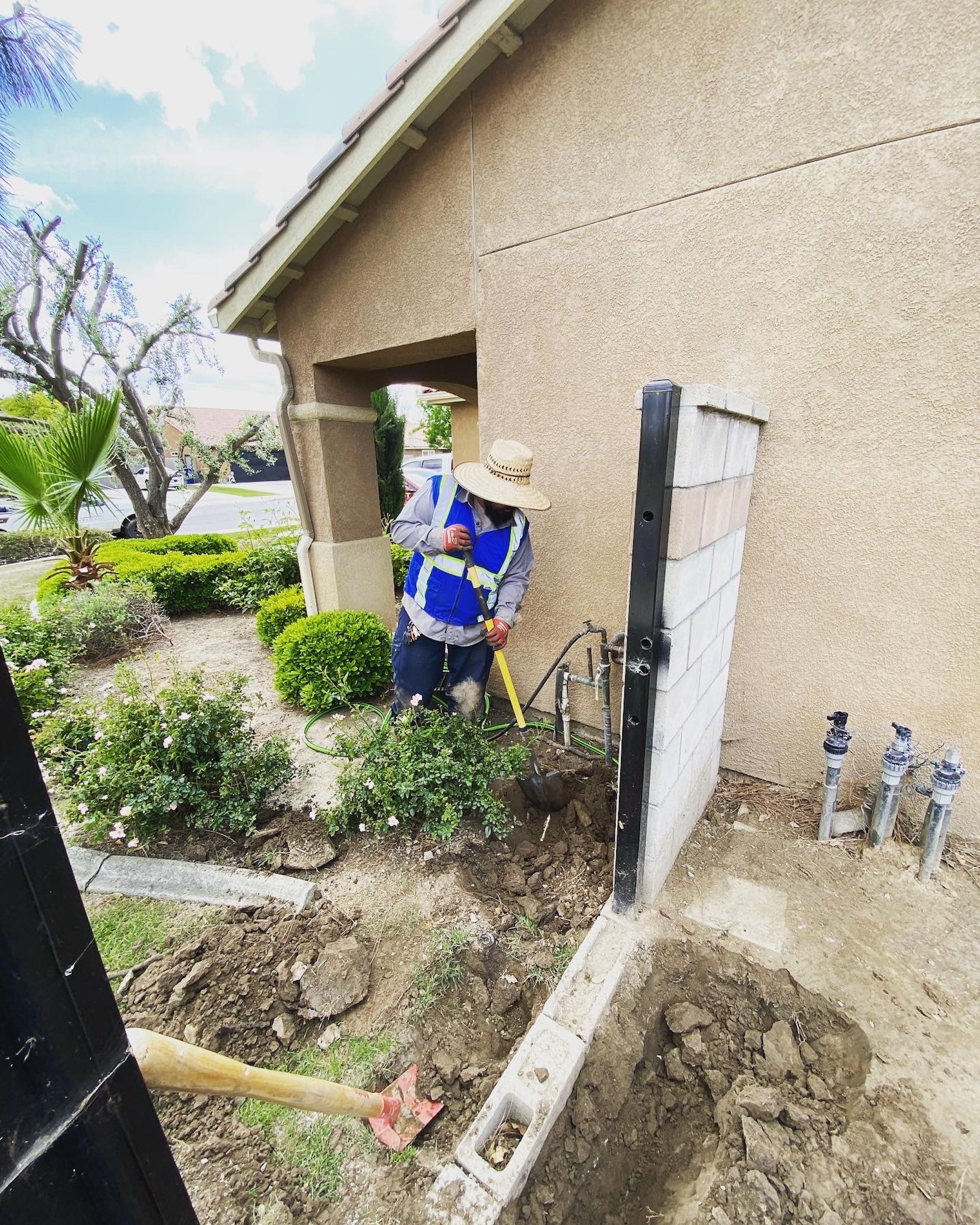 Worker digging near a stucco house and a concrete block wall, wearing a safety vest and hat.