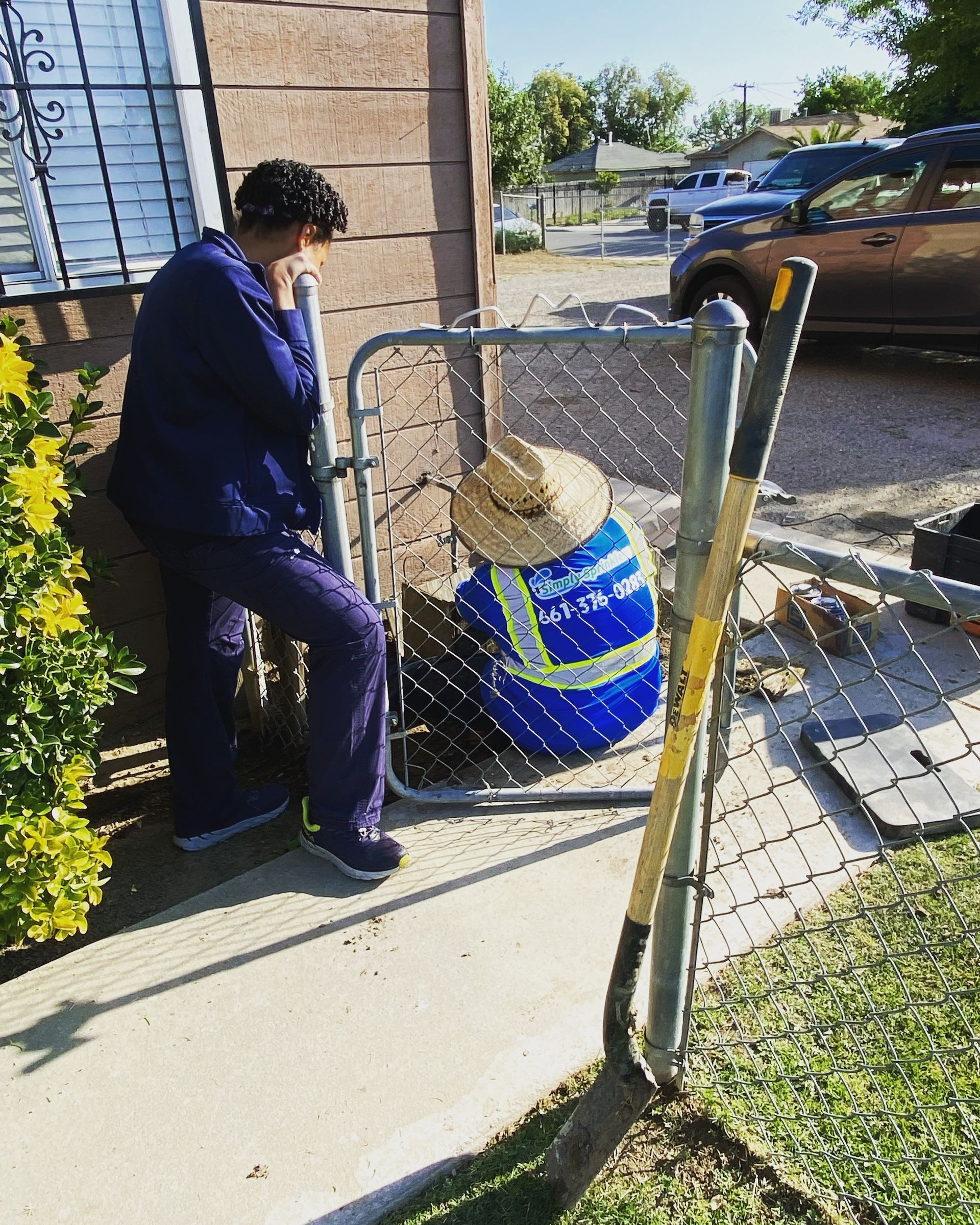 Person leans against a building, looking at another person behind a chain-link gate; a shovel is nearby.