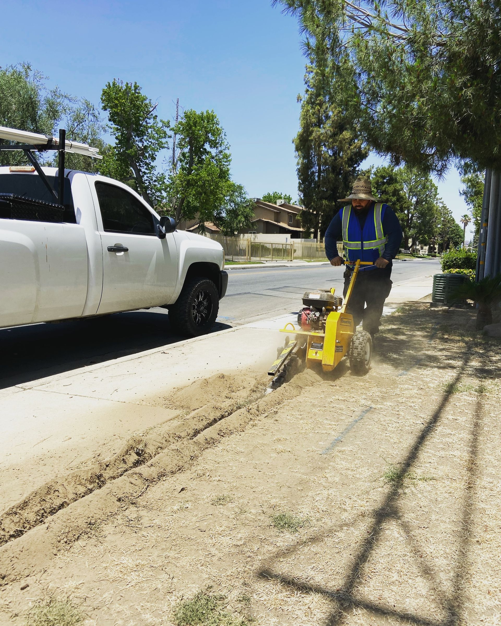 Man operating a small tiller on the side of a road, near a white truck and trees, on a sunny day.