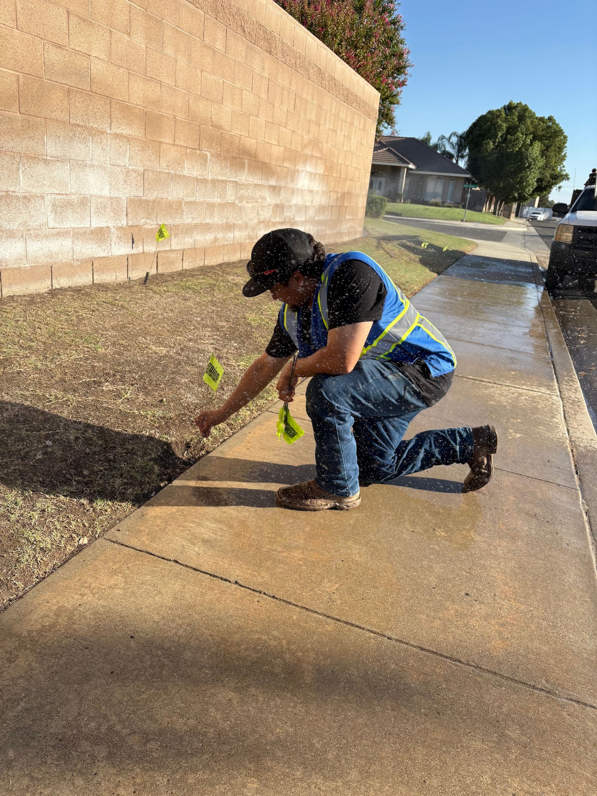 Person kneeling, adjusting sprinkler heads near sidewalk and beige wall, wearing reflective vest.