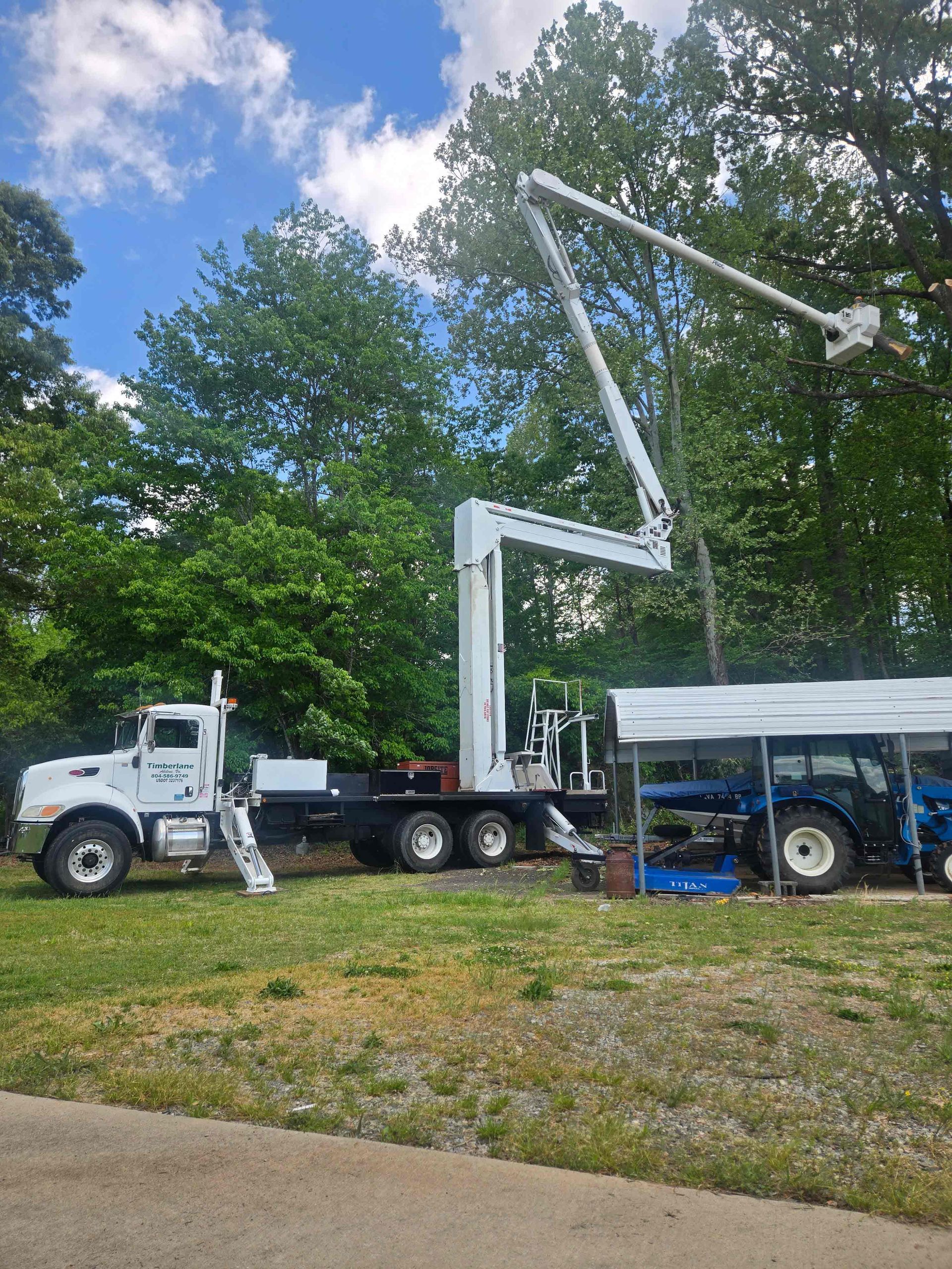 White utility truck with raised boom lift beside a covered trailer in a grassy lot under trees and blue sky